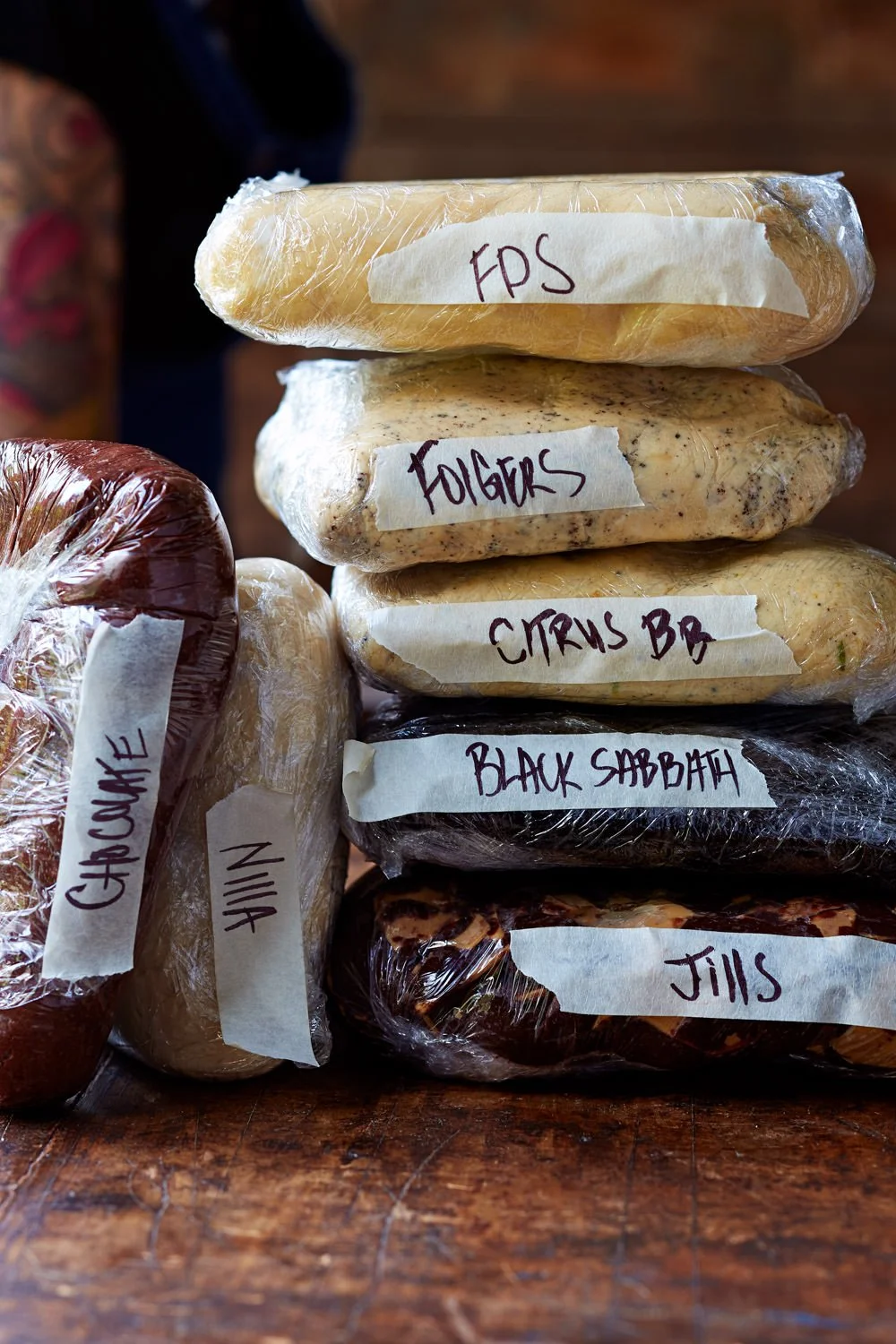Stacked homemade soap bars wrapped in plastic with handwritten labels: 'FDS', 'Folgers', 'Citrus Bar', 'Black Sabbath', 'Jills', 'ChowMe', and 'In' on a wooden surface.