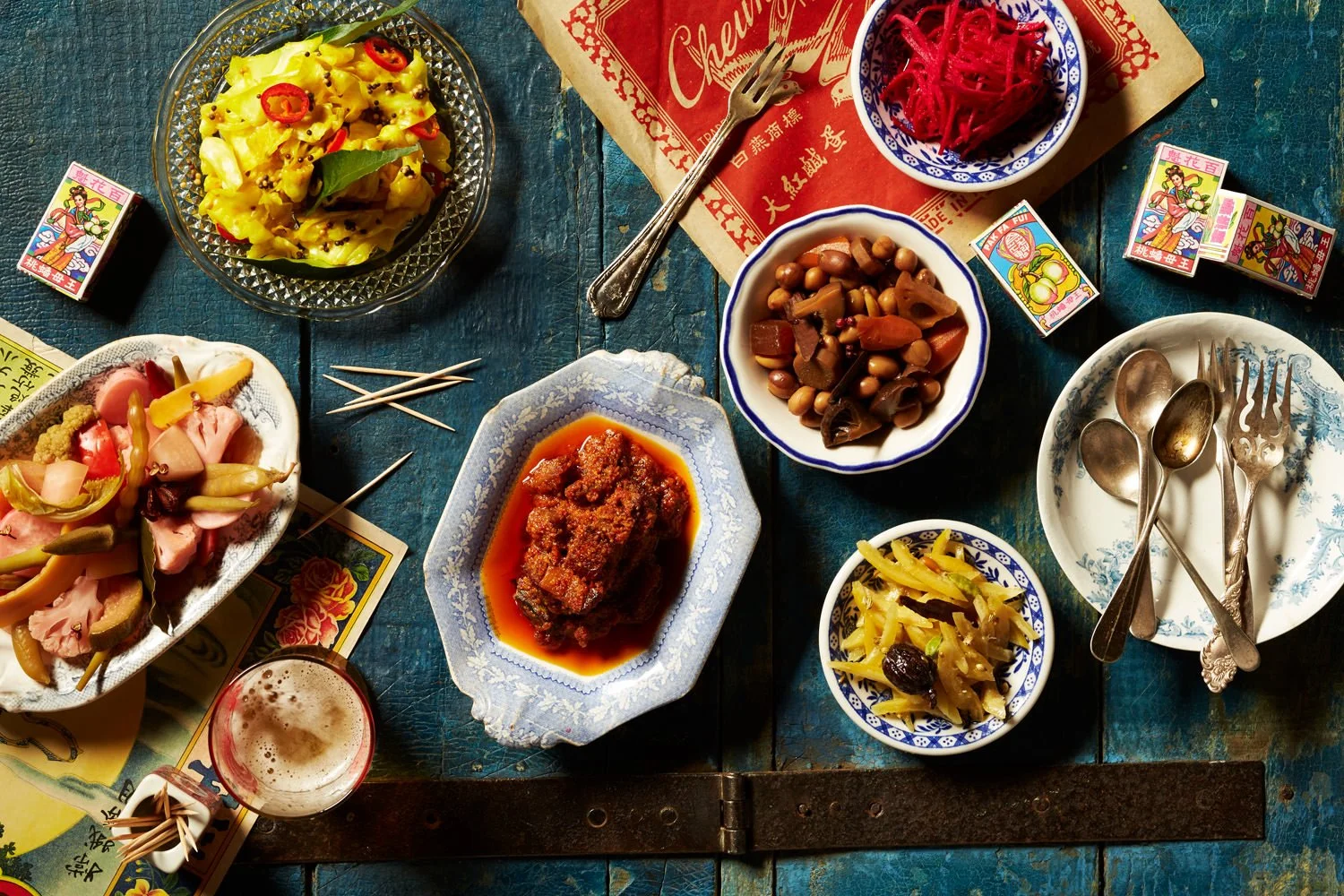 A table set with various Chinese dishes, small bowls of pickled vegetables, a glass of beer, and scattered playing cards.