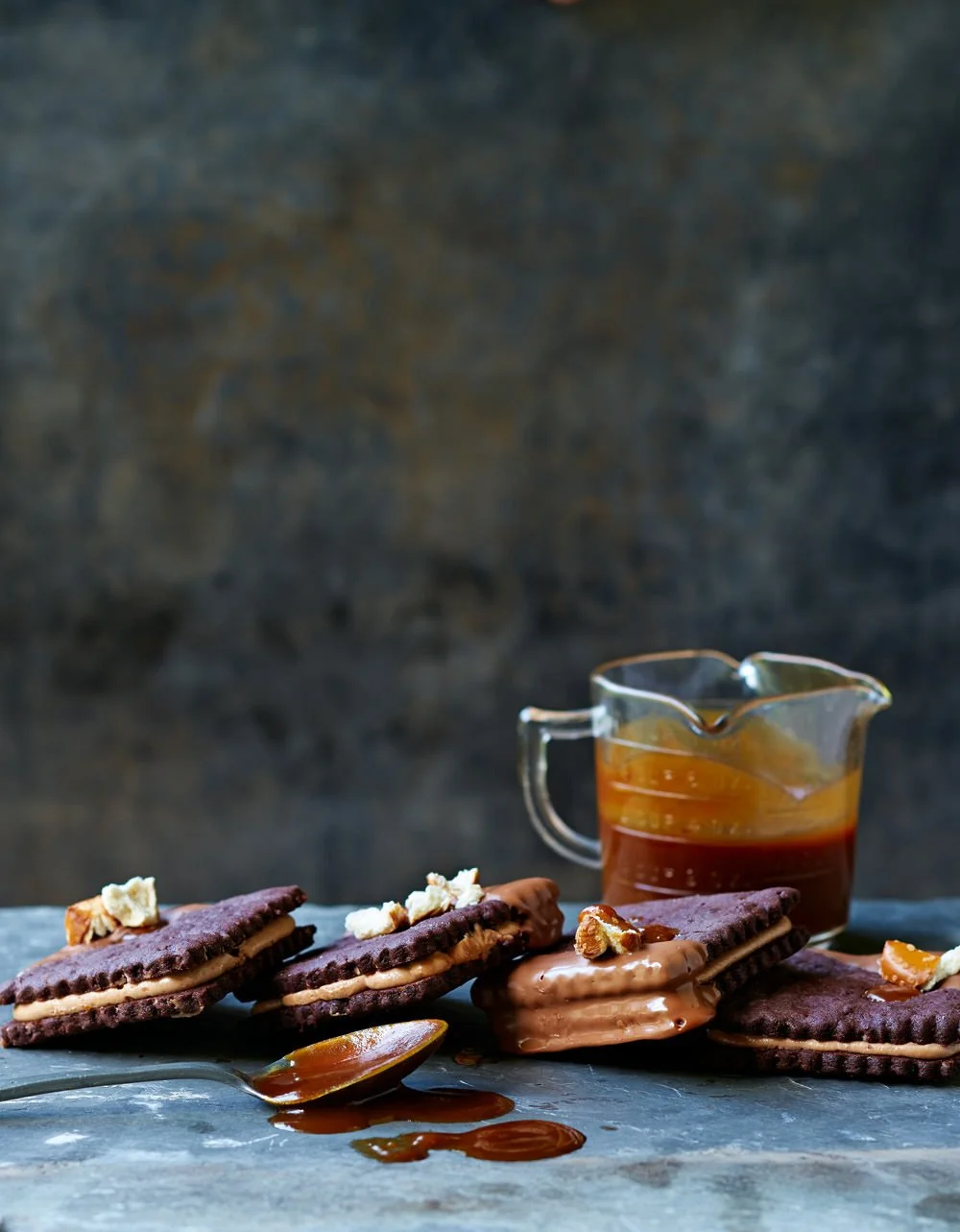 Chocolate cookies with caramel filling and walnuts, a glass pitcher of caramel sauce, and a spoon with caramel sauce on a gray surface against a dark background.