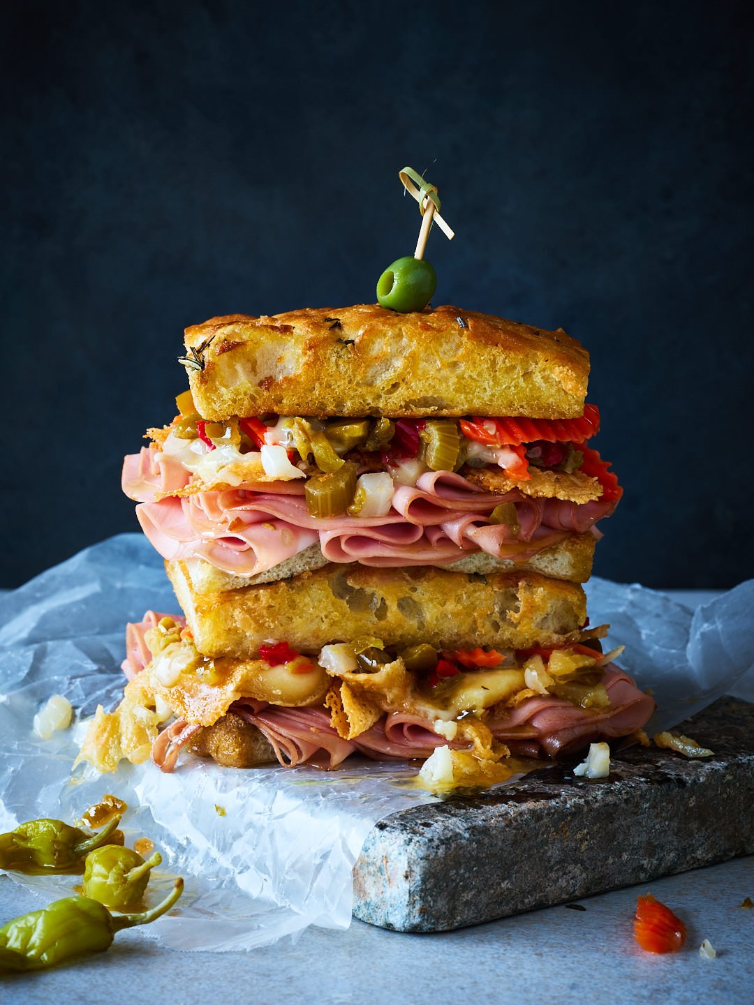 Stacked sandwich with breaded fried bread slices, ham, cheese, pickles, and vegetables, garnished with an olive pierced by a toothpick on top, placed on a stone slab lined with parchment paper.