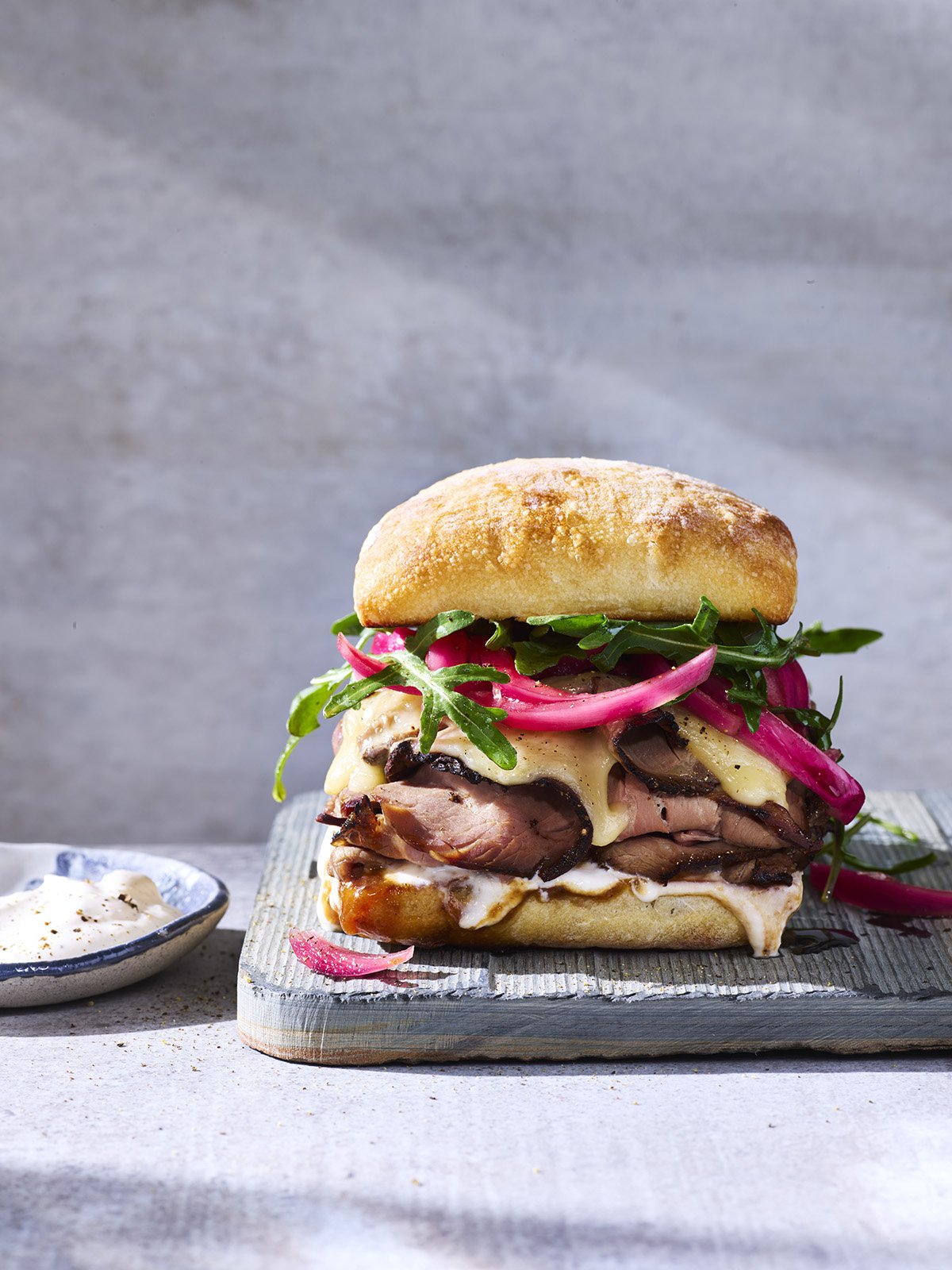A gourmet sandwich with roast beef, melted cheese, arugula, pickled red onions, on a bread roll, served on a wooden board with a side of mayonnaise or aioli.