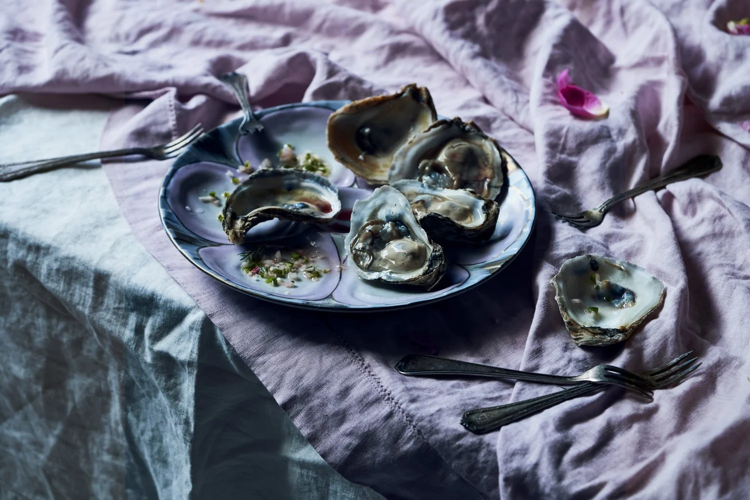 A plate of opened oysters with some green garnish on a pink cloth with several forks around it.