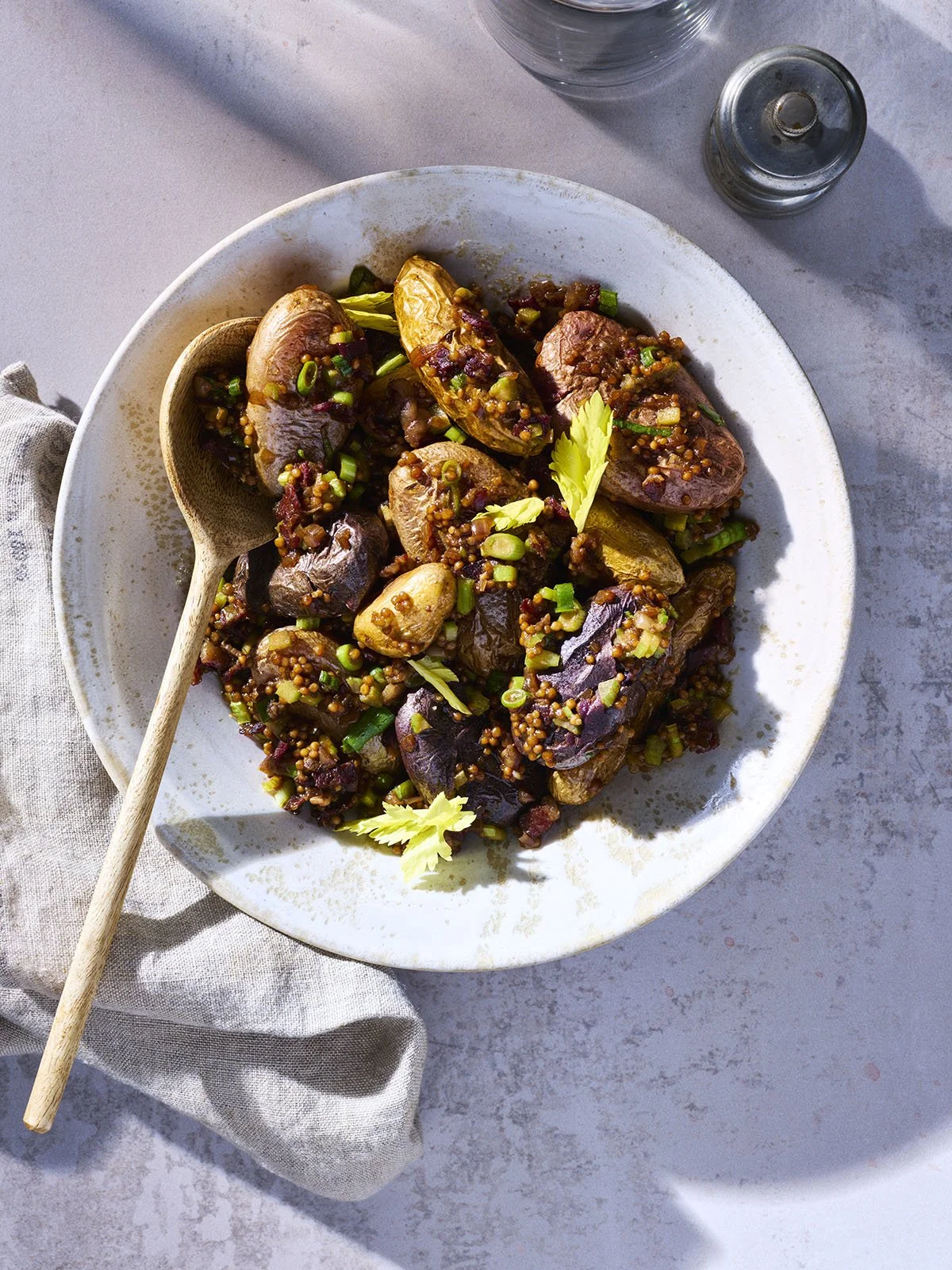 A plate of cooked eggplant with a sauce, garnished with chopped green onions and celery leaves, on a white table with a spoon and a glass of water.