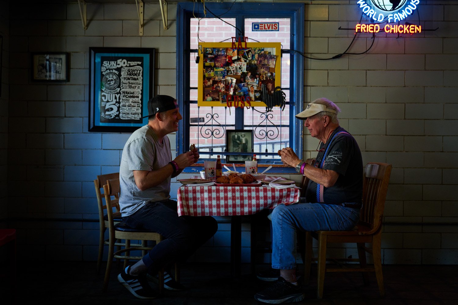 Two men sit across from each other at a small, round table with a red and white checkered tablecloth, enjoying fried chicken and drinks inside a restaurant. 