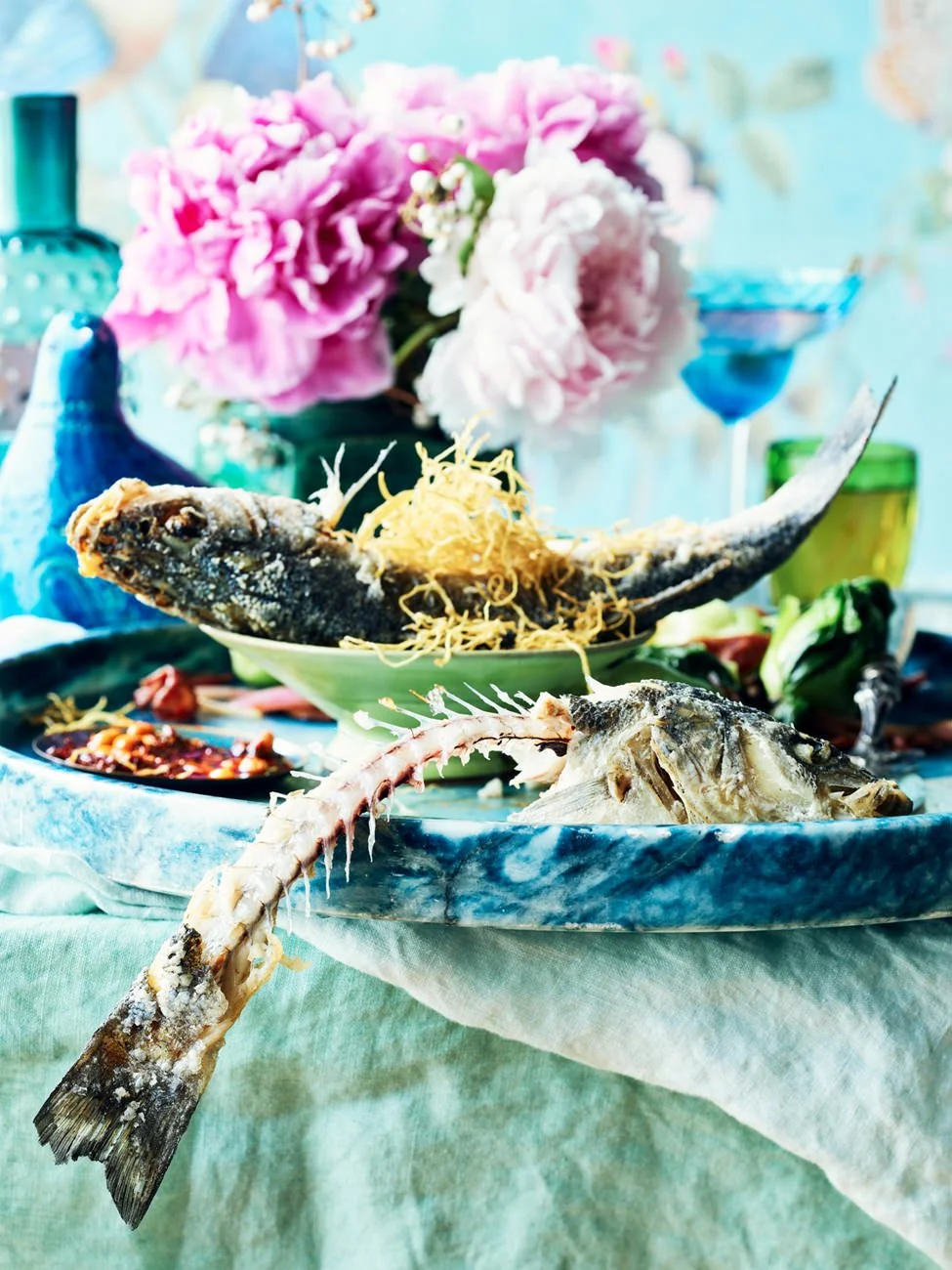 A table with a seafood dish featuring a whole cooked fish, fish head, and a fish tail on a marble platter, garnished with noodles. In the background are pink and white flowers, and colorful glassware.