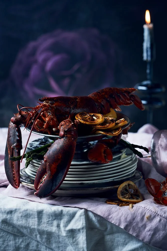 Lobster and oysters on a stack of plates with lemon slices, set on a table with a pink tablecloth, with a lit candle in the background.