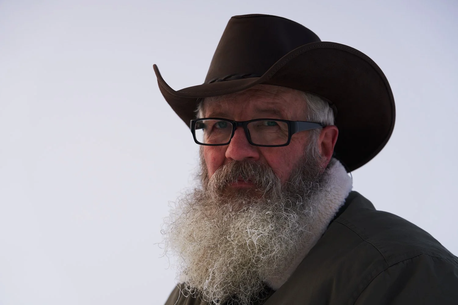 A man with glasses and a long white beard wearing a brown cowboy hat and a dark jacket, looking to the side against a plain background.