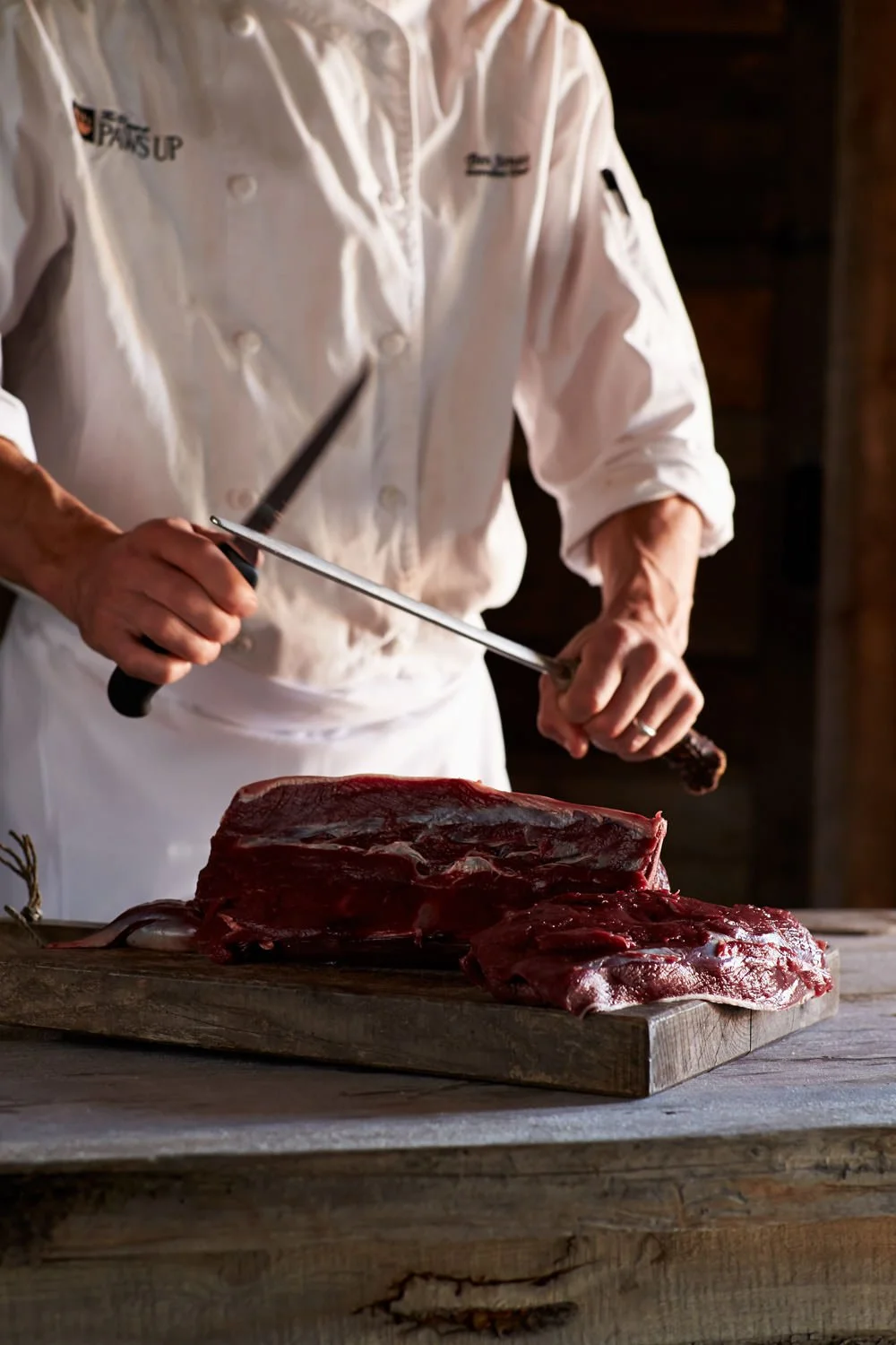 Chef in white uniform preparing to cut a large piece of raw beef on a wooden cutting board.