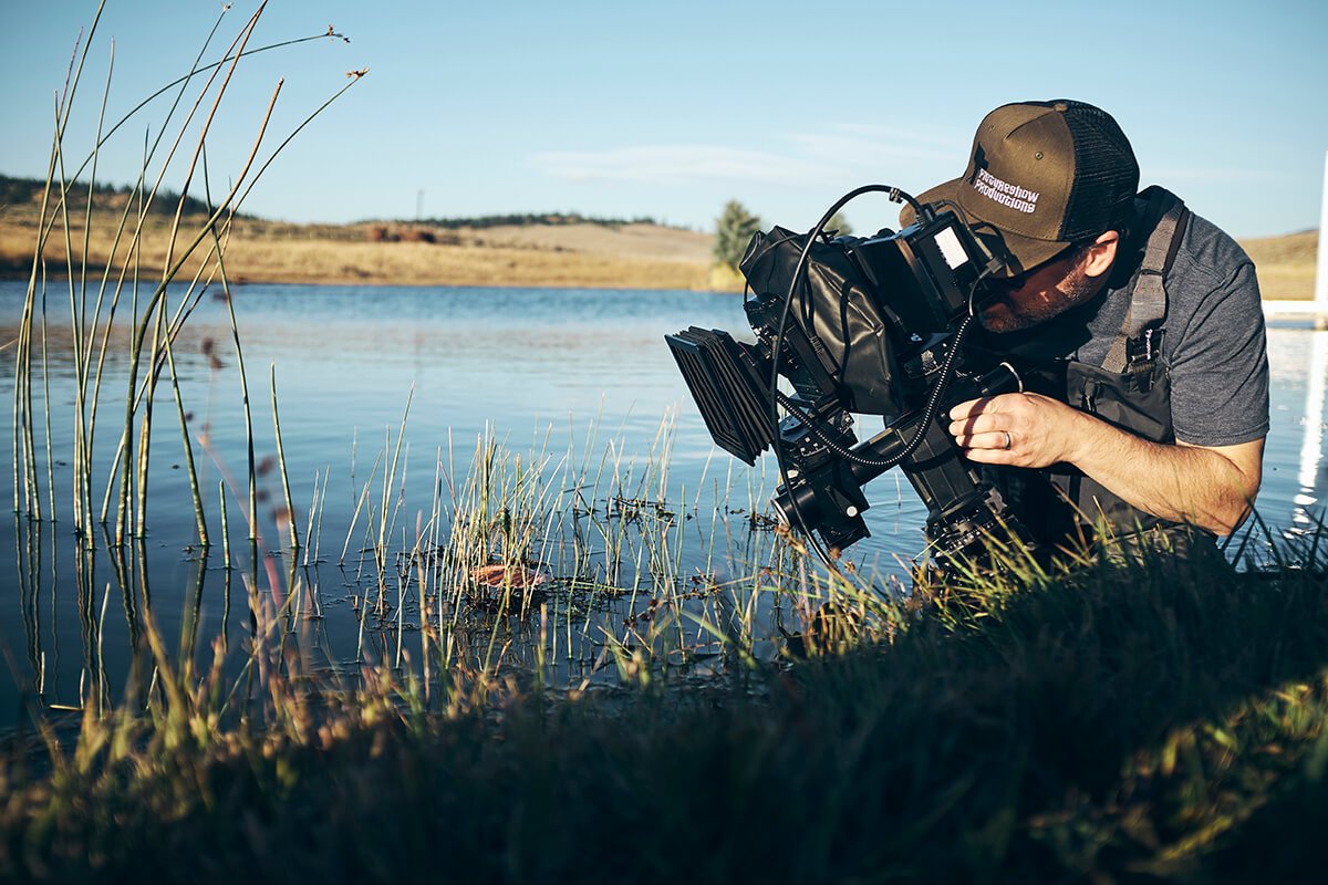 A photographer kneeling by a lake, adjusting a camera with a large lens, wearing a cap and outdoor gear.