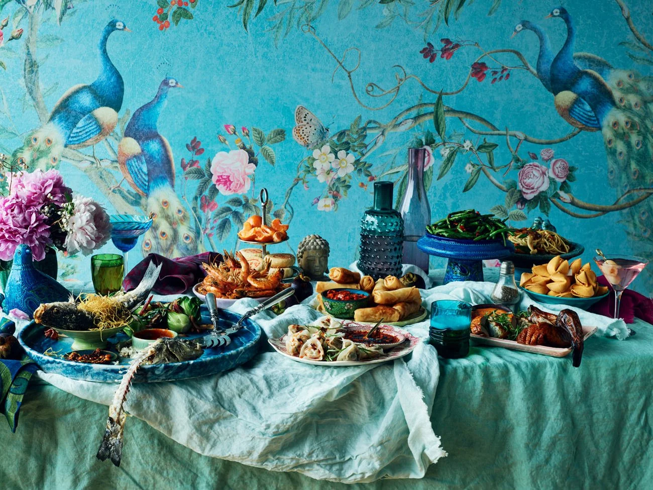 A colorful table set with various Asian dishes, fish, fried rolls, dumplings, vegetables, and sauces, with decorative vases and a peacock-themed wall in the background.