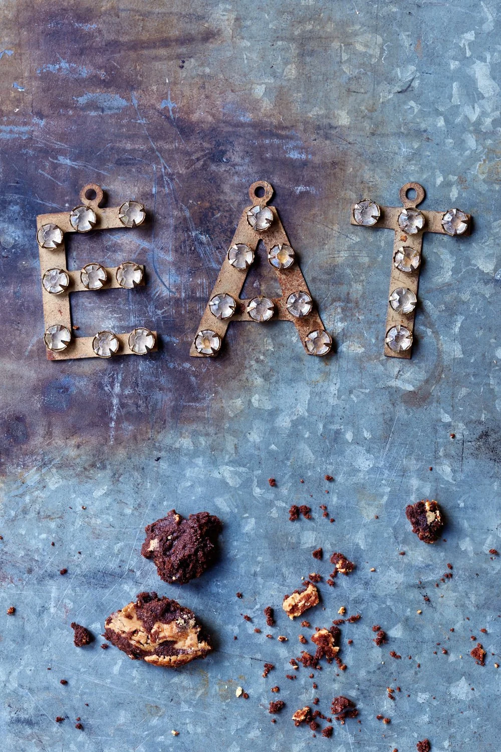 Metal letters spelling 'EAT' adorned with clear gemstones on a textured metal surface, with broken cookies and crumbs scattered below.