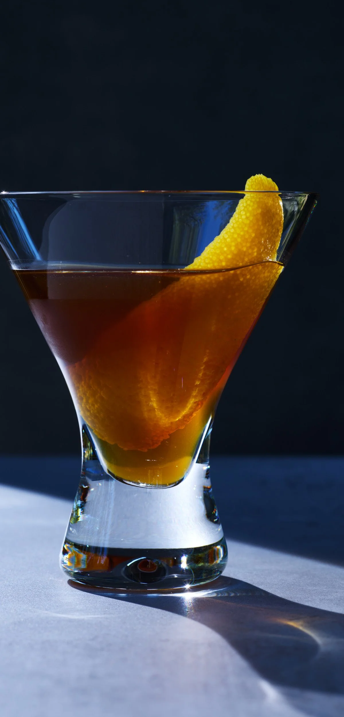 A cocktail in a coupe glass with an orange peel garnish, set on a white surface with a dark background.