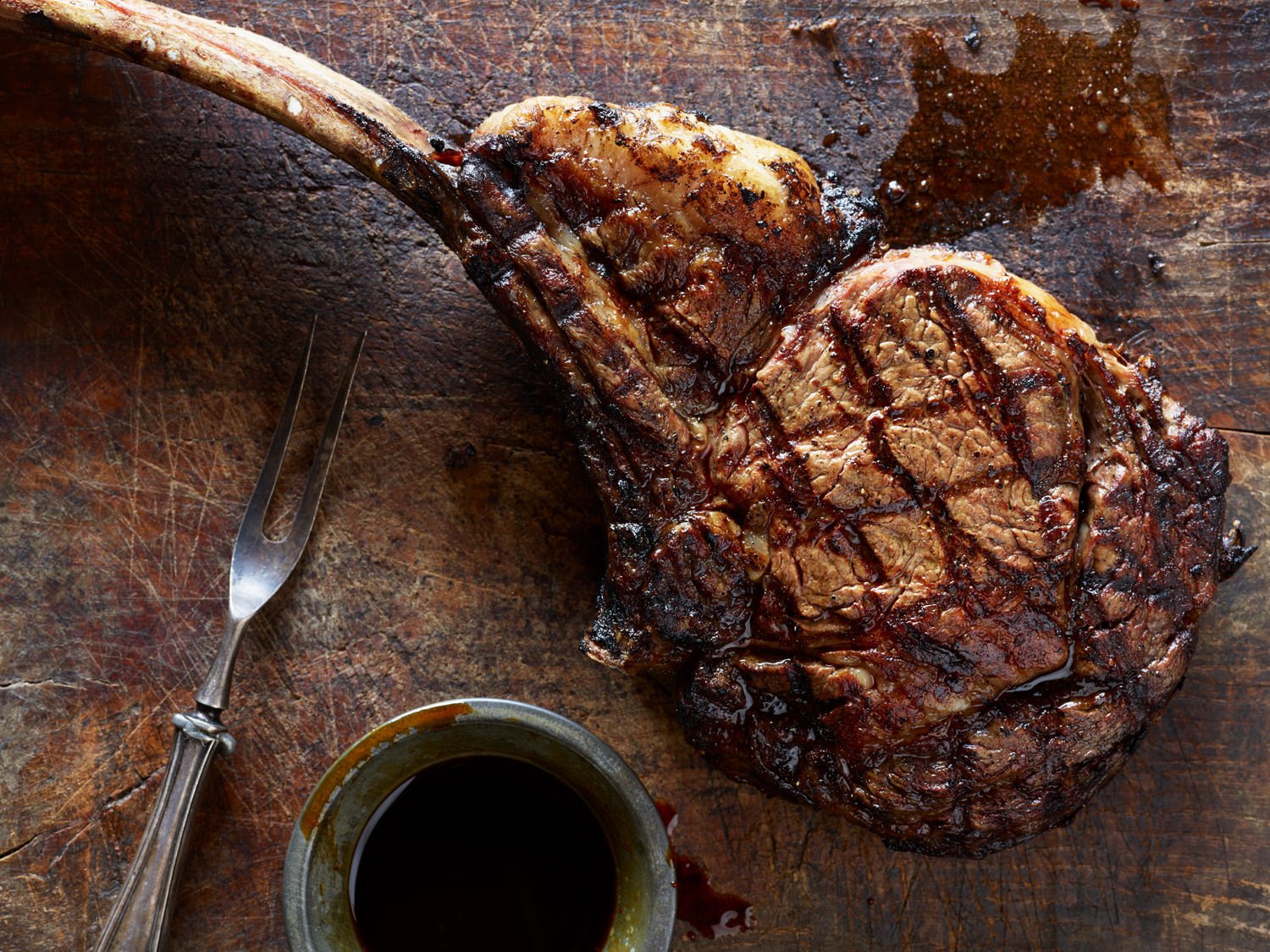 A large, cooked pork chop with visible grill marks on a wooden surface, next to a small bowl of dark barbecue sauce and a metal fork.