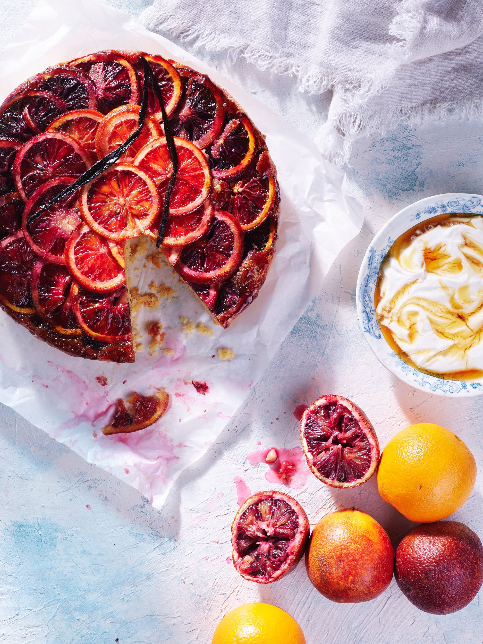 Blood orange upside-down cake with sliced blood oranges on top, a bowl of whipped cream or frosting, and whole blood oranges on a white textured surface.