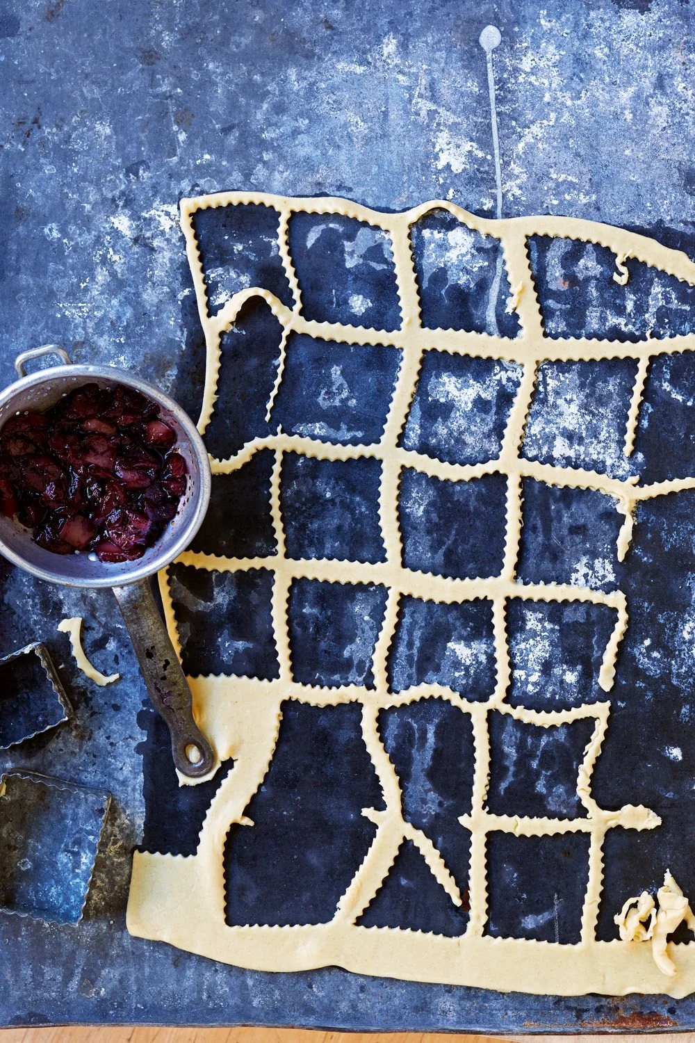 Uncooked pasta cut into a grid pattern on a metal surface with a bowl of cherry tomatoes and a metal cookie cutter nearby.