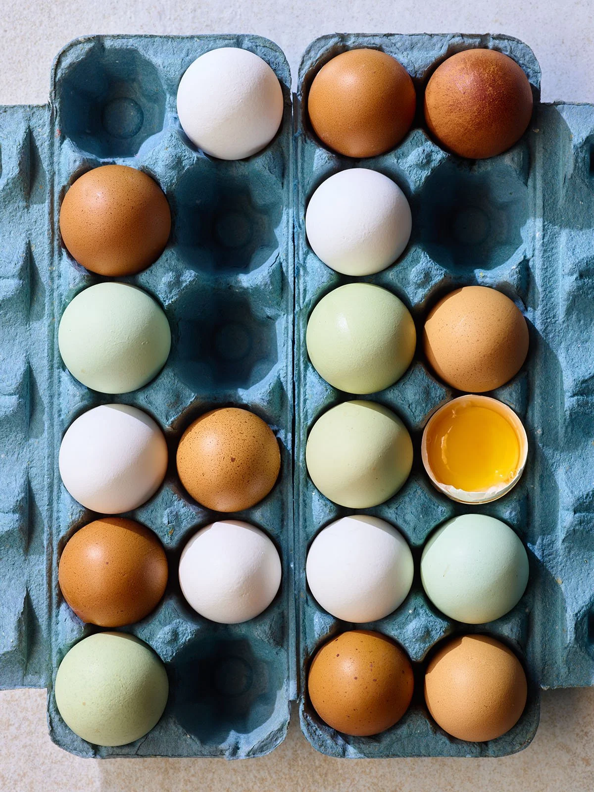 Various eggs, including white, brown, green-tinted, and one broken with visible yolk, arranged in a blue egg carton.