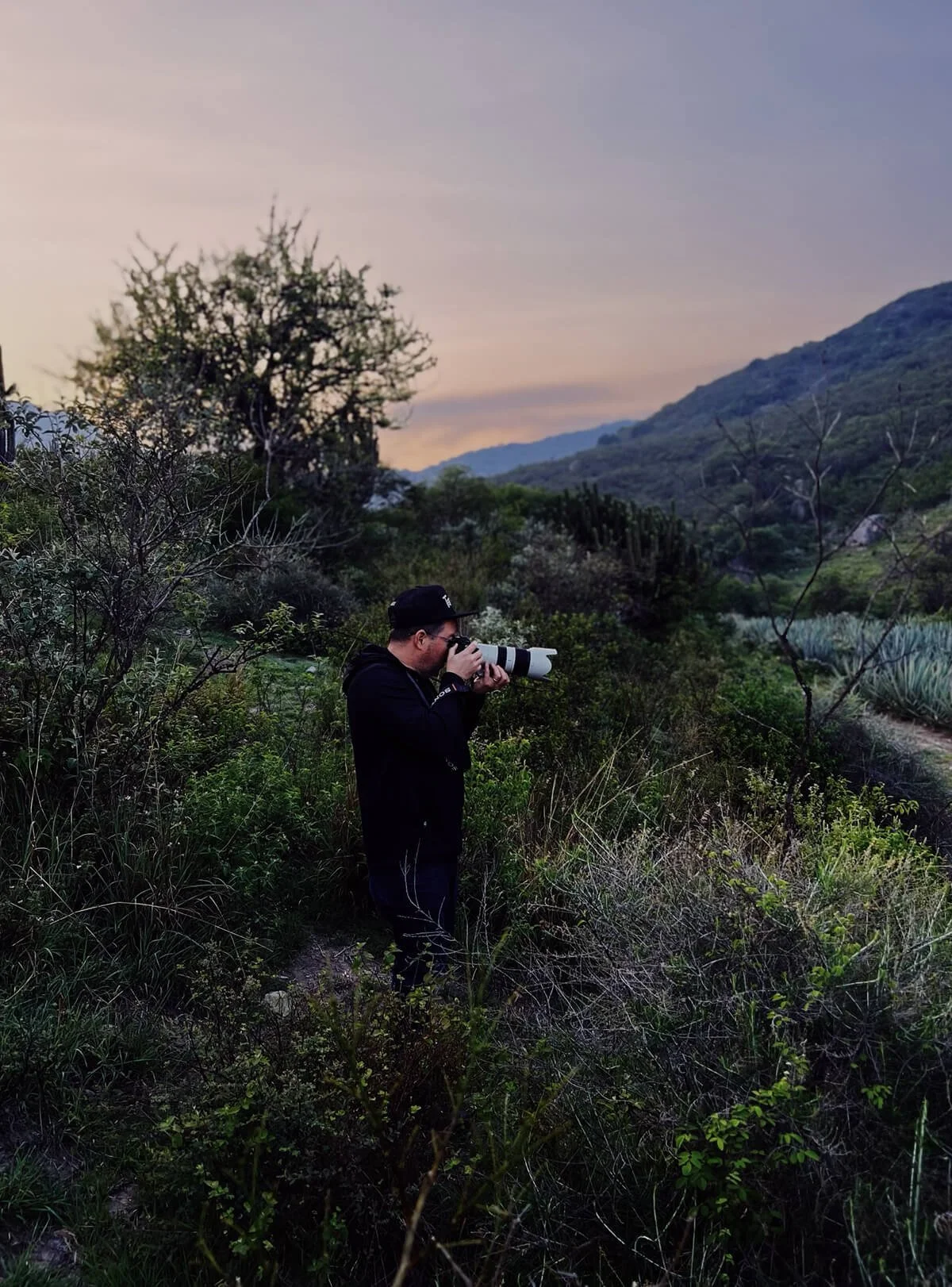Person with a camera in a green, hilly outdoor area during sunset.
