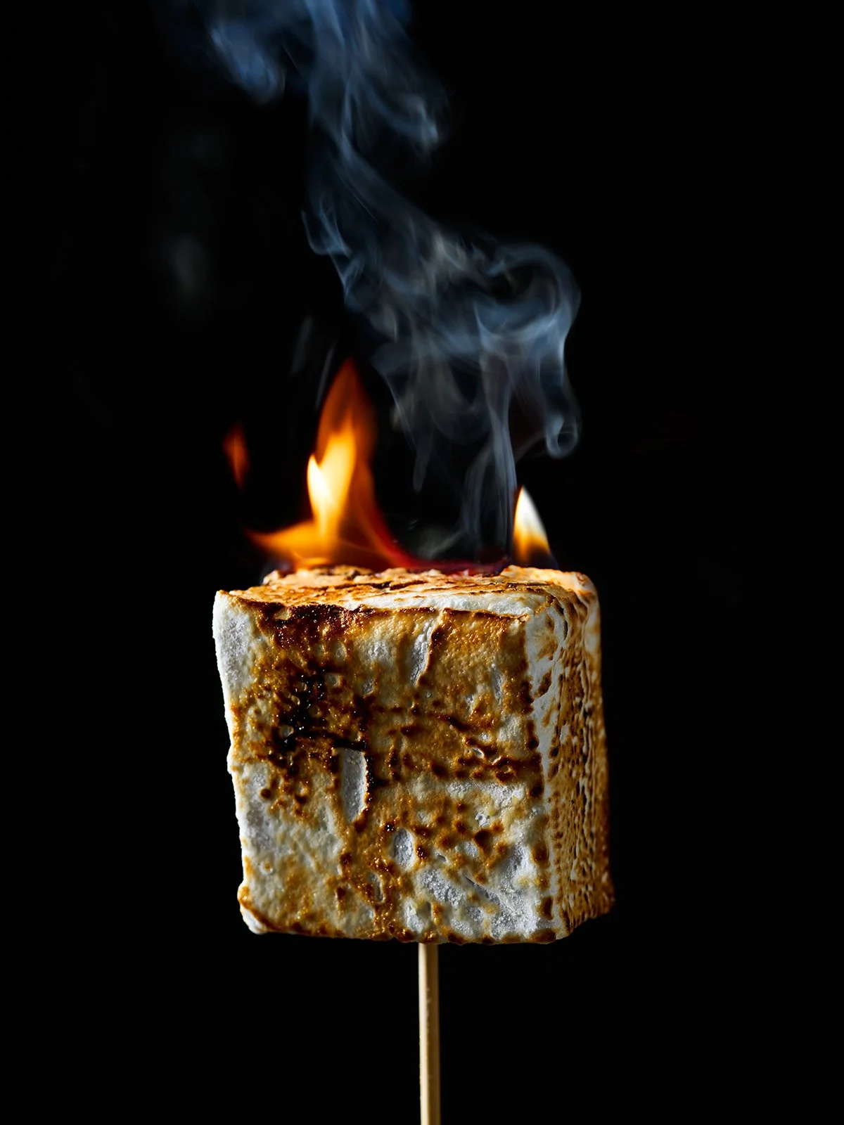 A toasted marshmallow on a stick burning with flames and smoke on a black background.