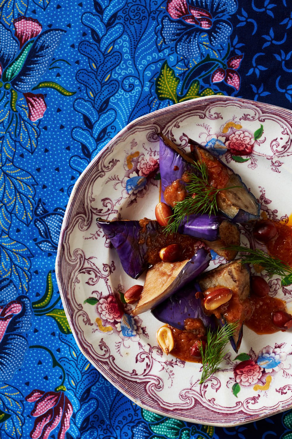 Close-up of a floral-patterned porcelain plate with cooked eggplant, garnished with herbs, nuts, and tomato-based sauce, on a vibrant blue floral fabric background.