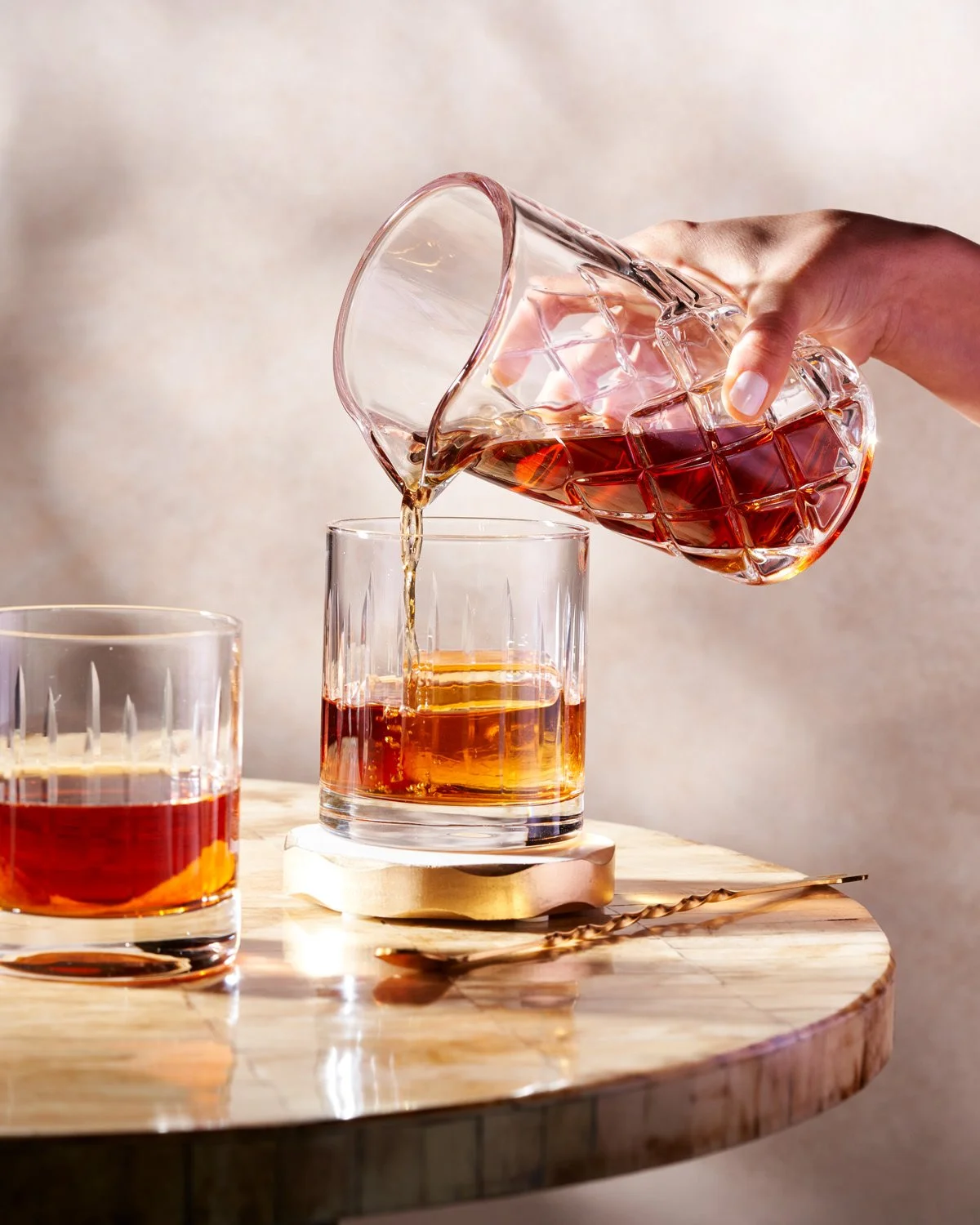 A person pouring whiskey from a glass pitcher into a tumbler glass on a wooden surface, with another glass of whiskey nearby.