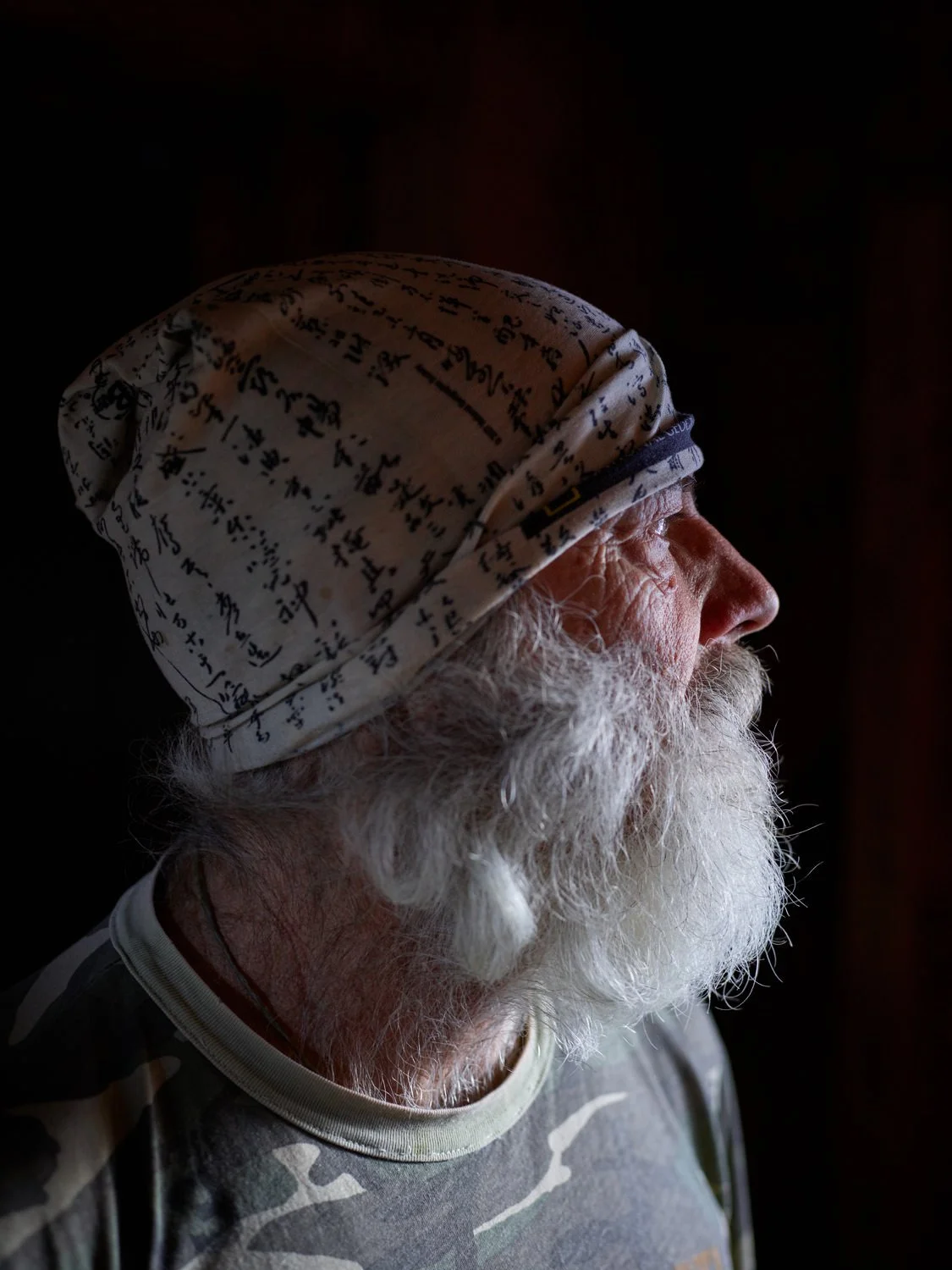 Side profile of an elderly man with a white beard and mustache, wearing a beanie with handwritten text, and a camouflage-patterned shirt, in a dark background.