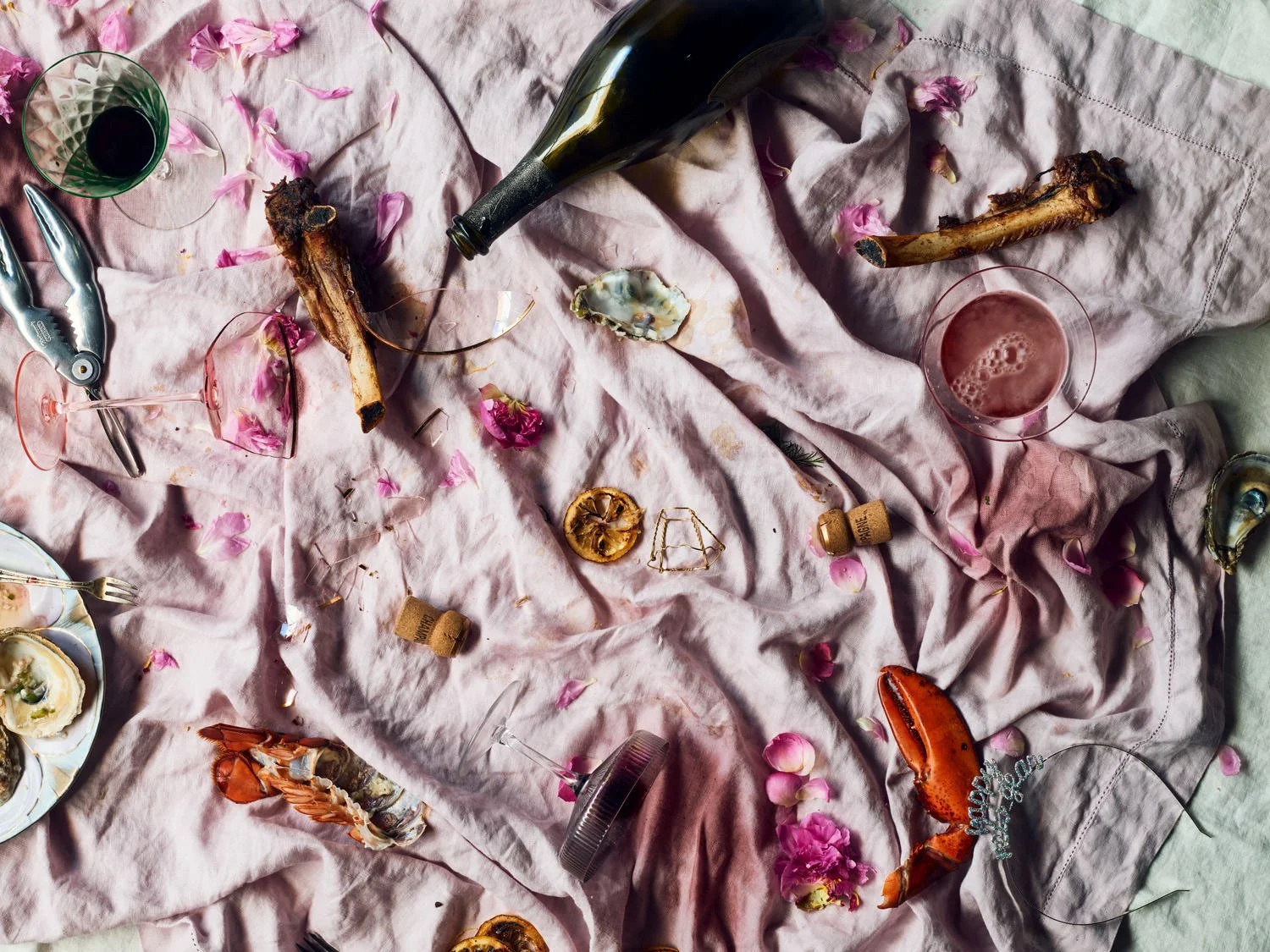 Messy table with shells, wine bottles, glassware, lobster claws, dried lemon slices, and rose petals on a pink and gray cloth.