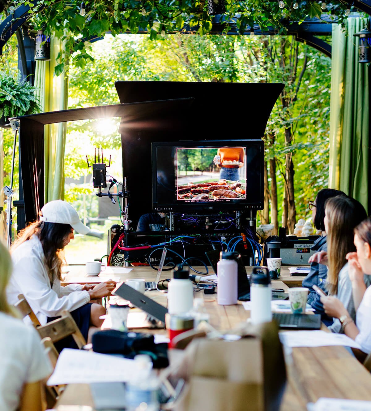 A group of people on a film set outdoors around a long table with laptops and notes, with a large camera and monitor capturing a scene of grilling food outdoors, surrounded by green trees and plants.