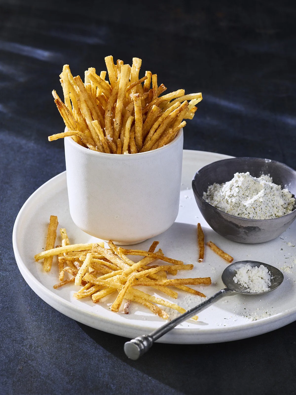 French fries in a white cup, with additional fries on a white plate, a small black bowl of flour, and a metal spoon with flour on a white plate.