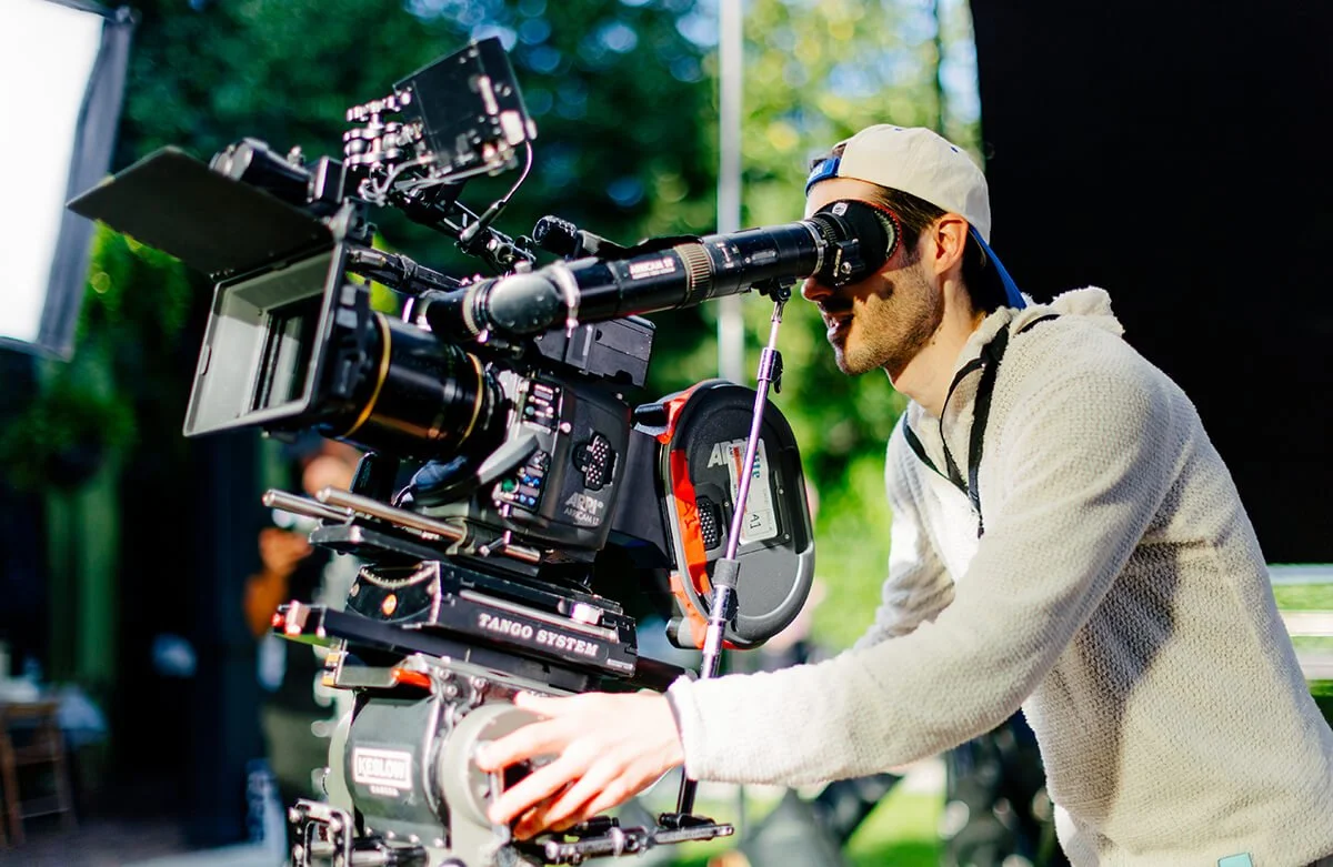 Filmmaker operating a professional camera on a set outdoors with trees in the background.