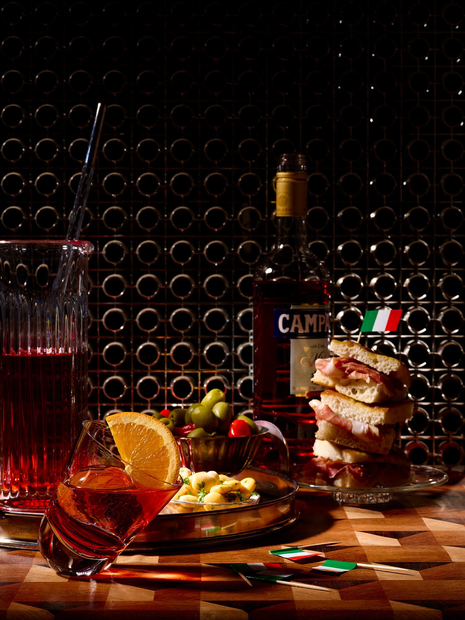 An Italian-themed bar setting with a bottle of Campari, a glass of Campari with an orange slice, a pitcher of Campari with a straw, a small bowl of green and red grapes, a plate of pasta with white beans, and a sandwich with prosciutto on a glass pla