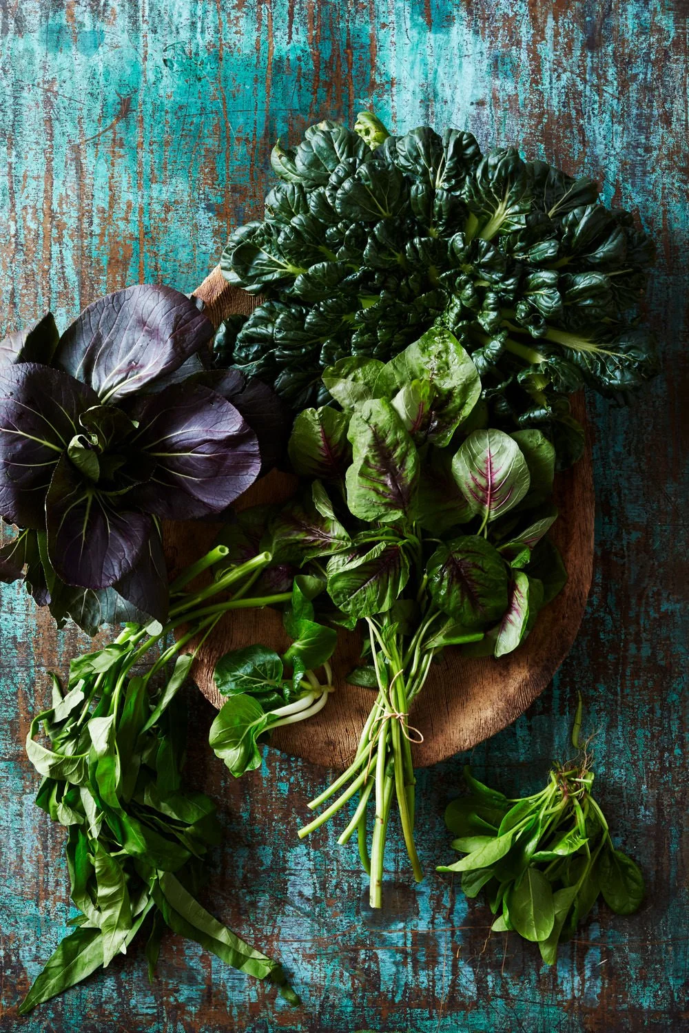 Assorted fresh leafy greens including spinach, chard, and purple perilla on a rustic wooden cutting board against a distressed blue wooden background.