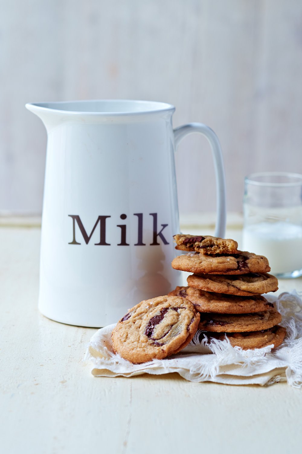 A white pitcher labeled 'Milk' beside a plate of chocolate chip cookies on a white cloth with a glass of milk in the background.