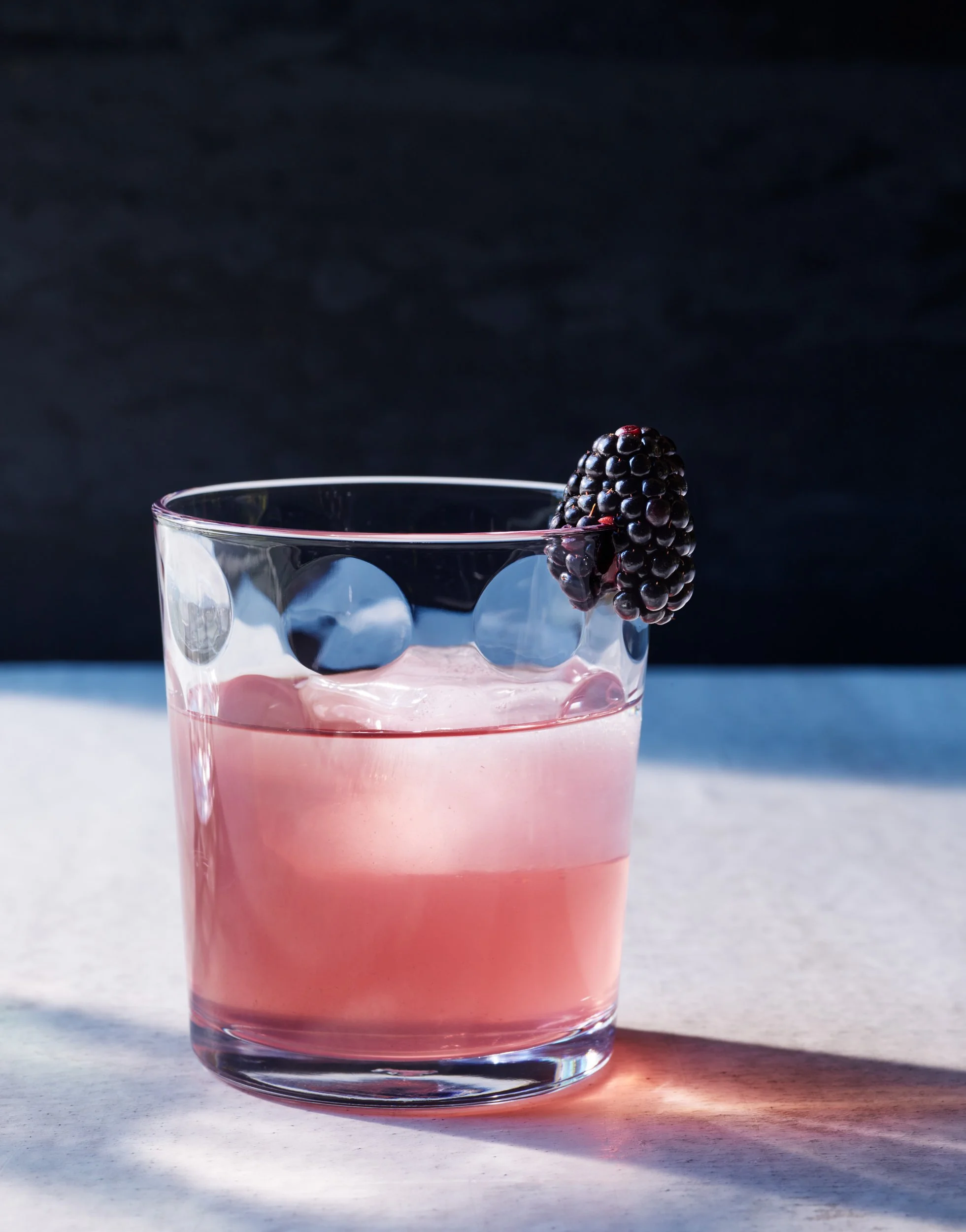 A glass of pink beverage garnished with a blackberry on the rim, set on a light-colored surface with a dark background.
