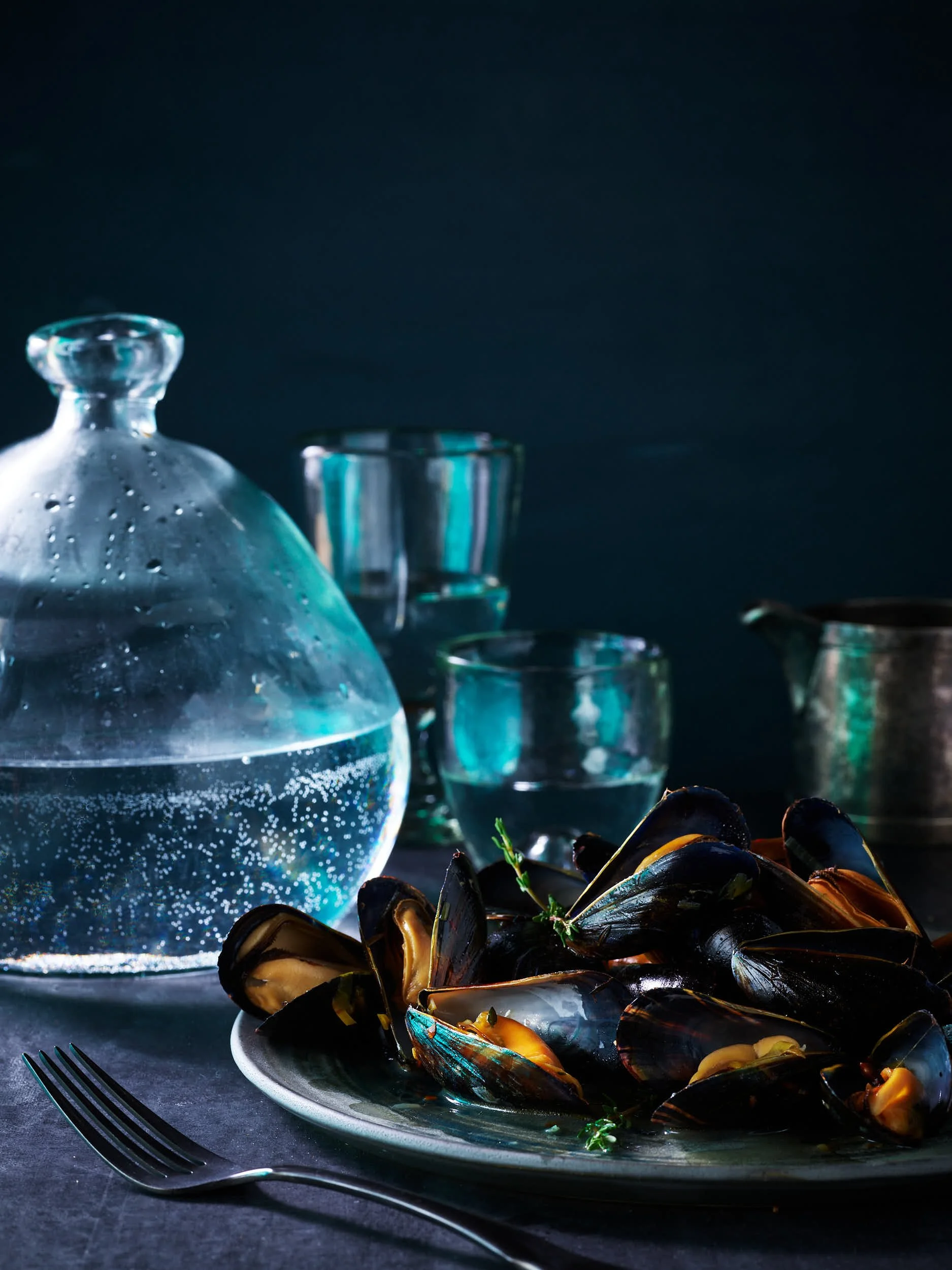 A plate of cooked mussels garnished with herbs on a dark table, with a glass decanter of water and drinking glasses in the background.