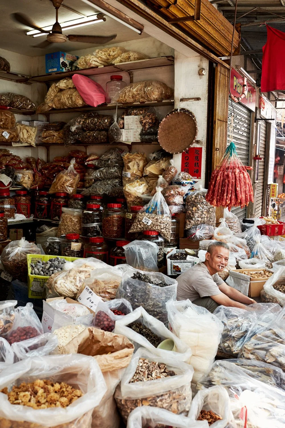 A man is sitting among various dried herbs, roots, and spices in bags at a market stall. The stall is filled with jars and bags of dried goods, with shelves above holding more spices and ingredients. There is a ceiling fan overhead and some hanging r