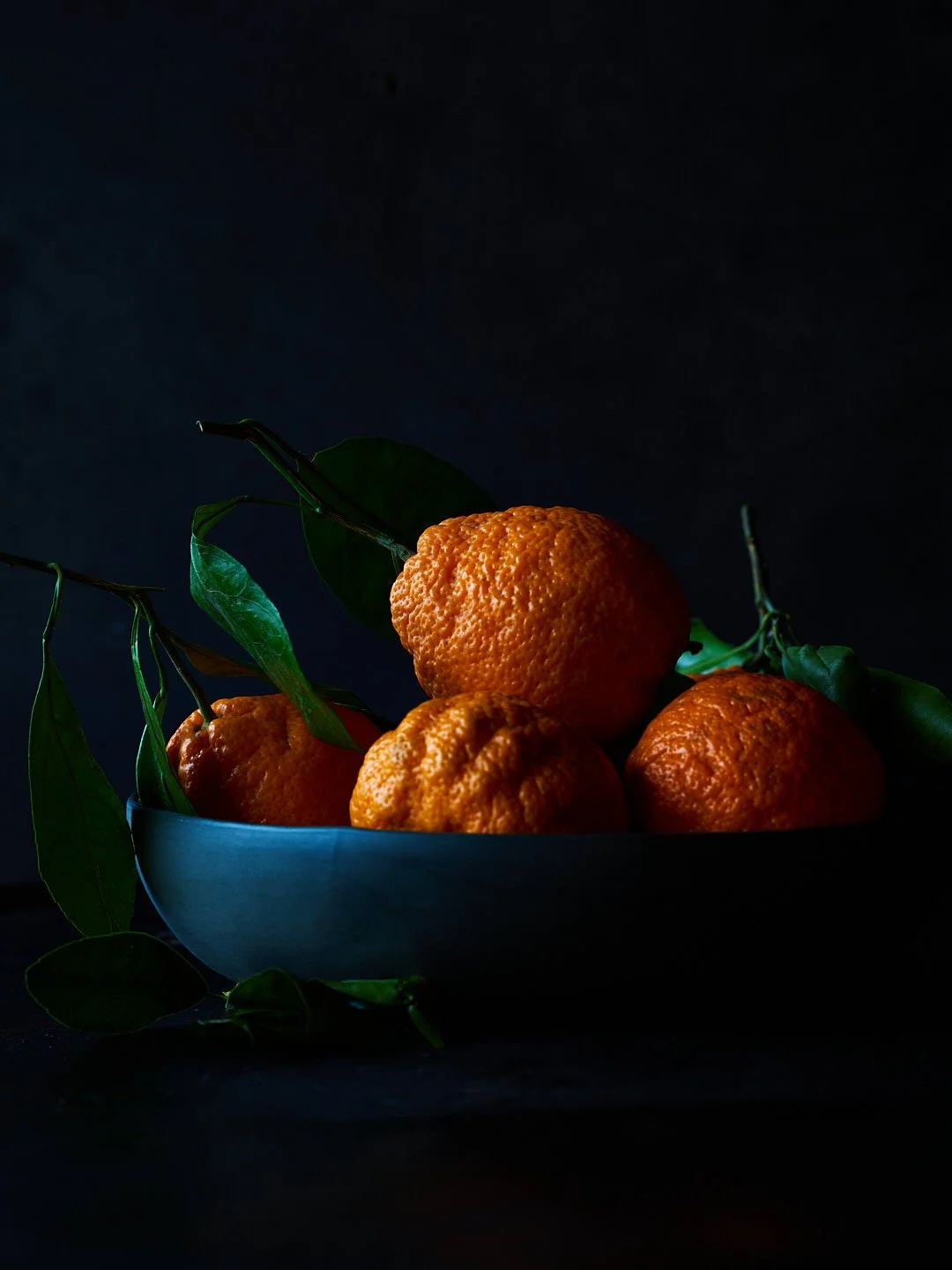 A black bowl filled with fresh, bright orange mandarins with green leaves still attached, set against a dark background.