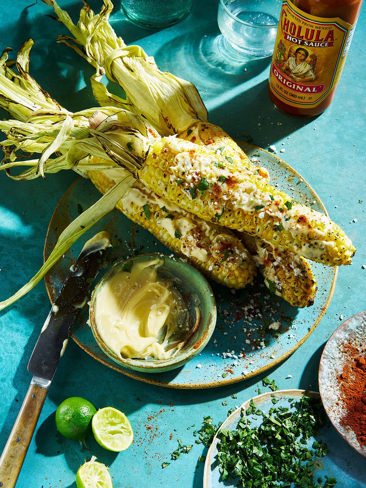 Grilled corn on the cob with some herbs and powder on top, a small bowl of butter, a sliced lime, a small bowl of hot sauce, and some chopped herbs on a blue table.