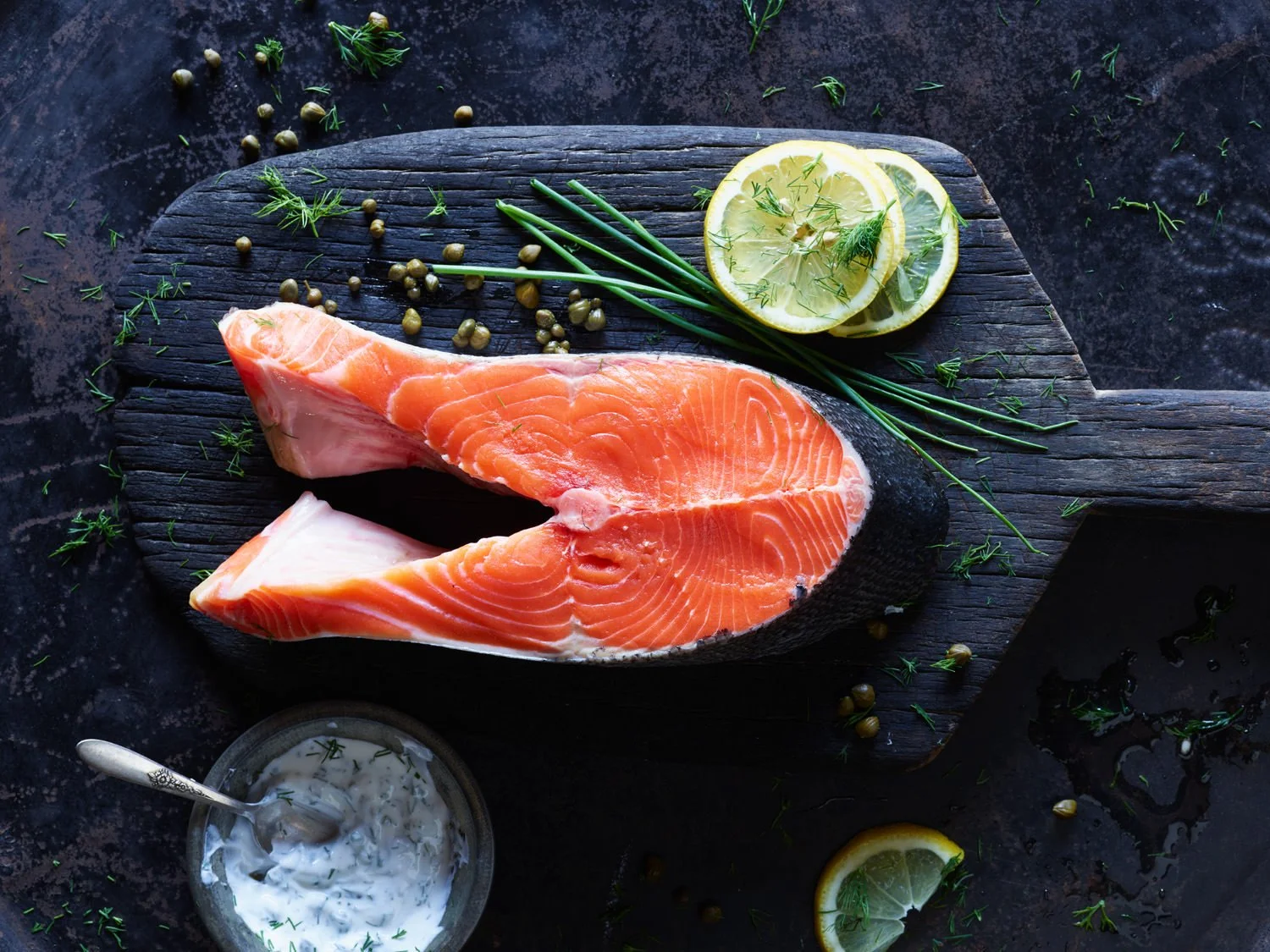 Raw salmon fillet on a dark wood cutting board with lemon slices, fresh herbs, and a small bowl of creamy sauce.