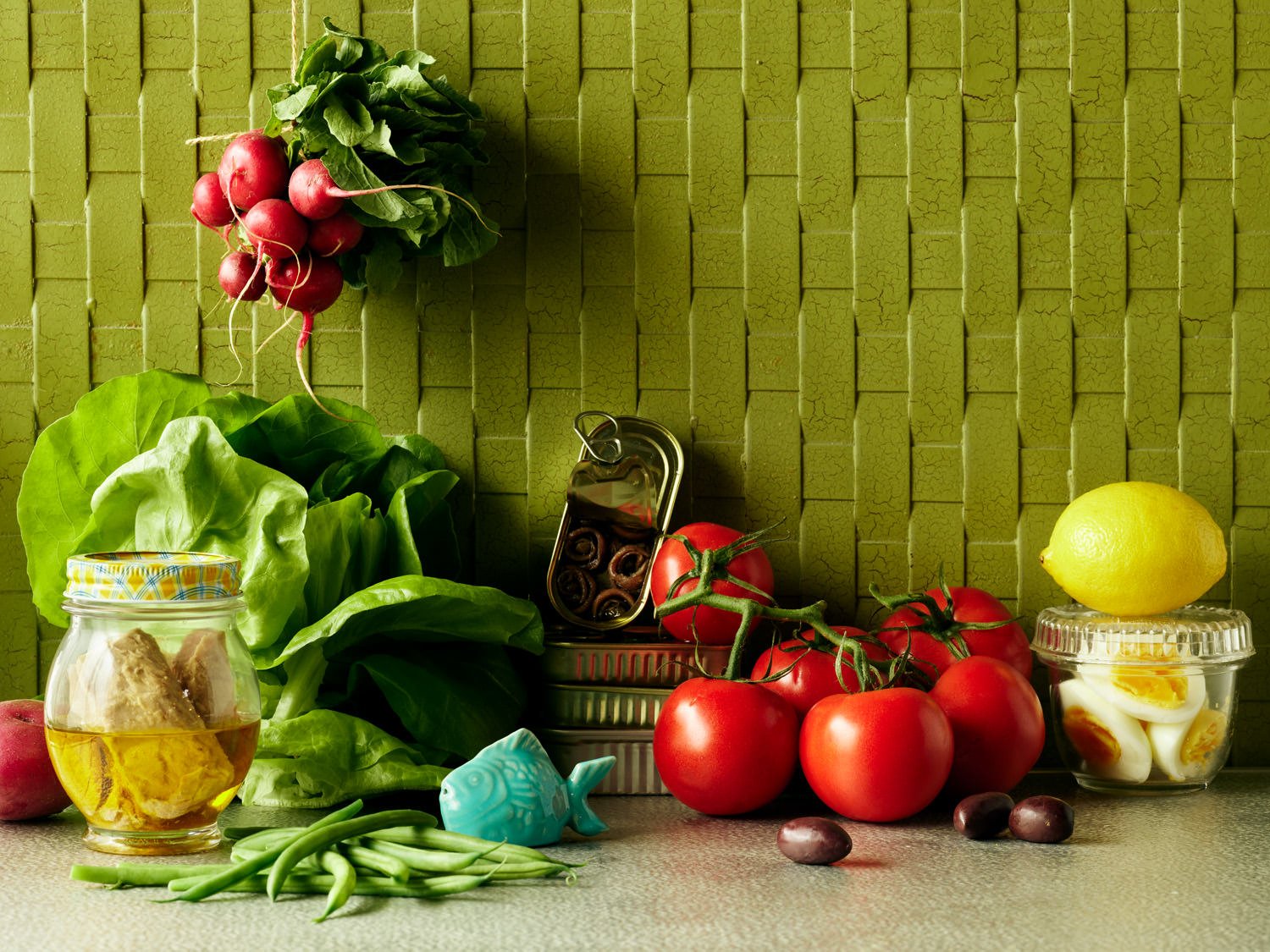 Fresh vegetables and food items on a kitchen countertop, including radishes, lettuce, tomatoes, lemon, hard-boiled eggs, grapes, and a jar of honey, with a textured green wall in the background.