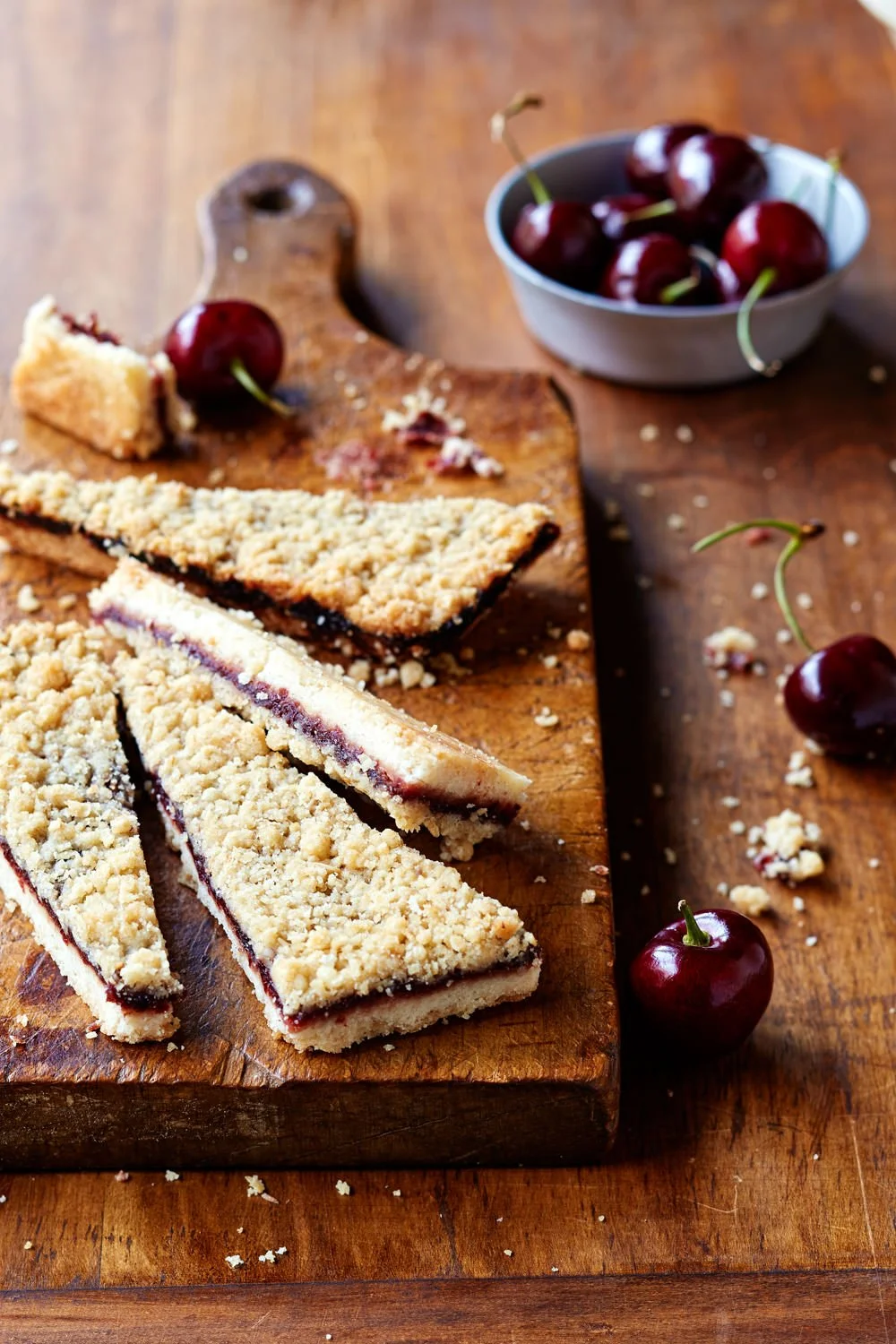 Slices of cherry crumble pie on a wooden cutting board with fresh cherries and a bowl of cherries in the background.