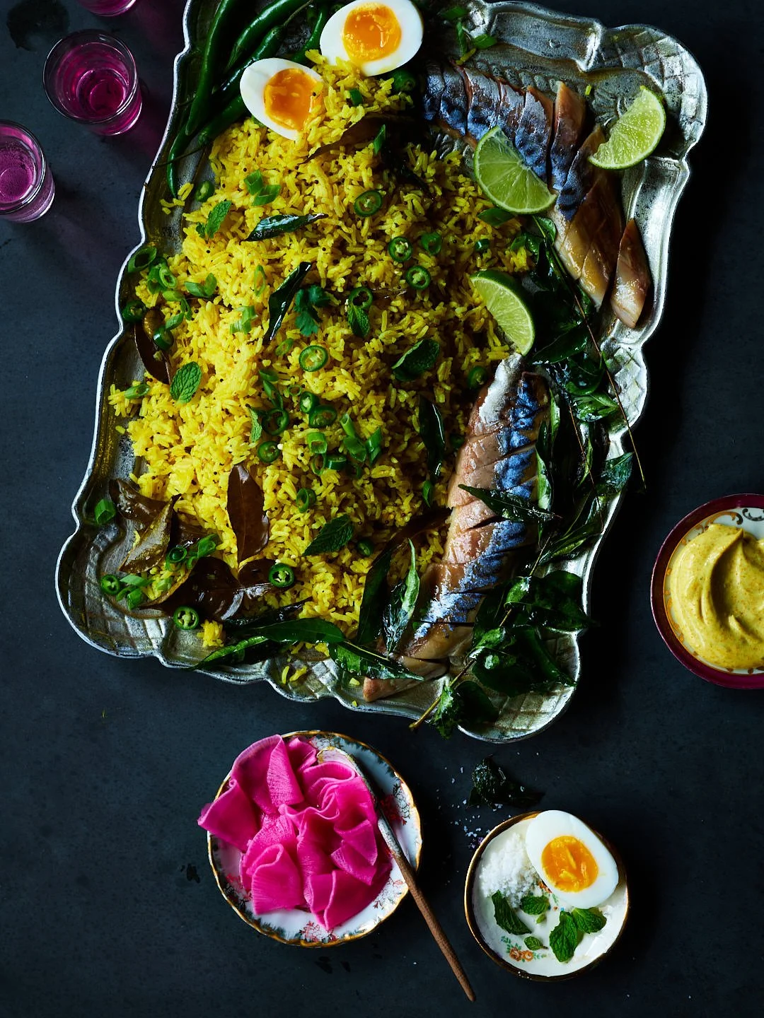 A large silver tray of saffron rice garnished with green chili slices, mint leaves, and bay leaves, accompanied by grilled fish with lemon wedges, halved boiled eggs, a bowl of yellow mustard, and a small bowl of pink pickled vegetables on a dark sur