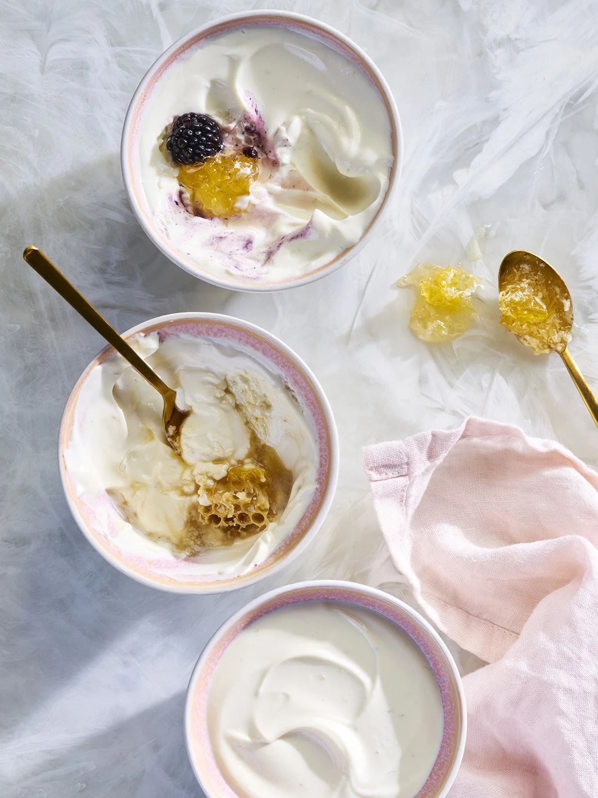 Three cups of yogurt with honey and mixed berries, placed on a marble surface with honey drizzles and a pink cloth.