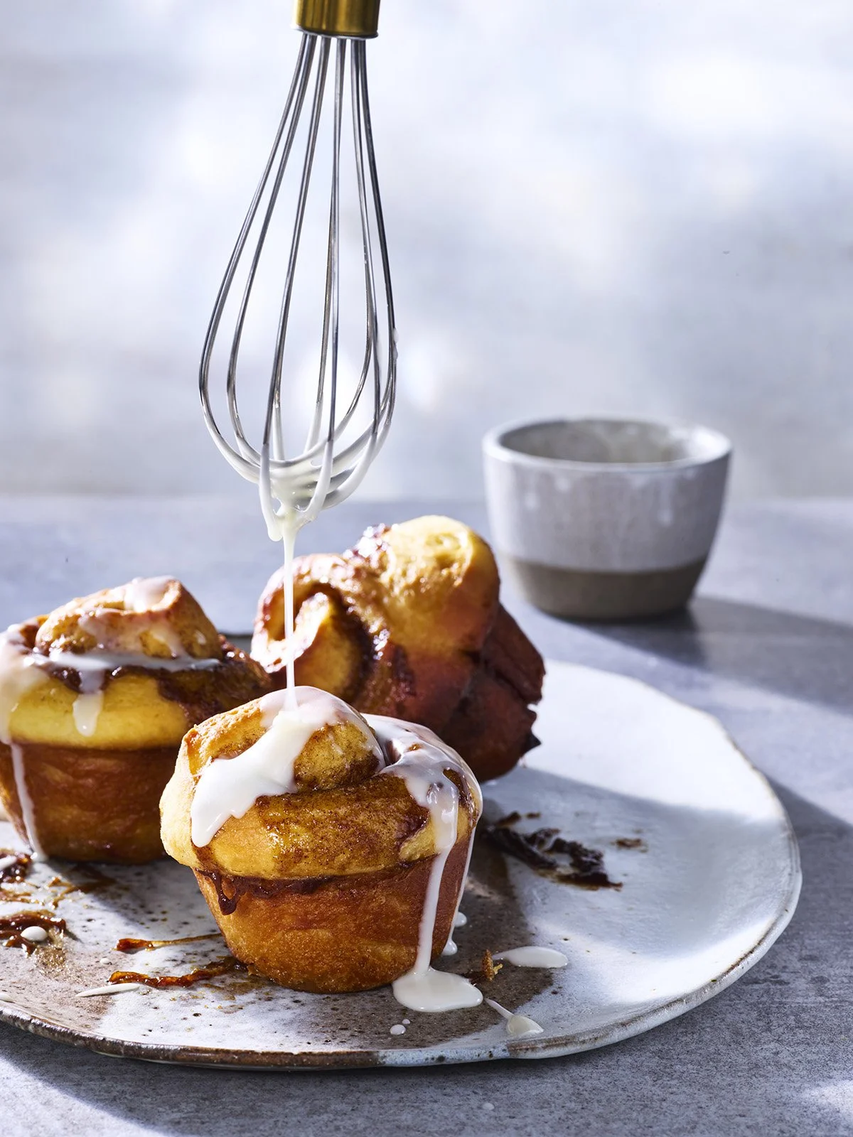 Cinnamon rolls topped with white icing on a ceramic plate, with a whisk drizzling icing over them and a small bowl in the background.