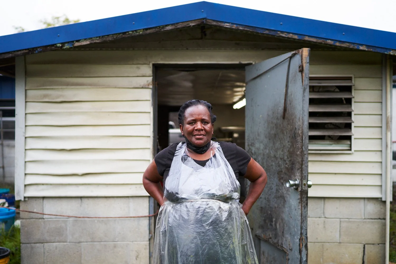 A woman standing outside a small, weathered house with open door, wearing a black shirt and a translucent plastic apron.