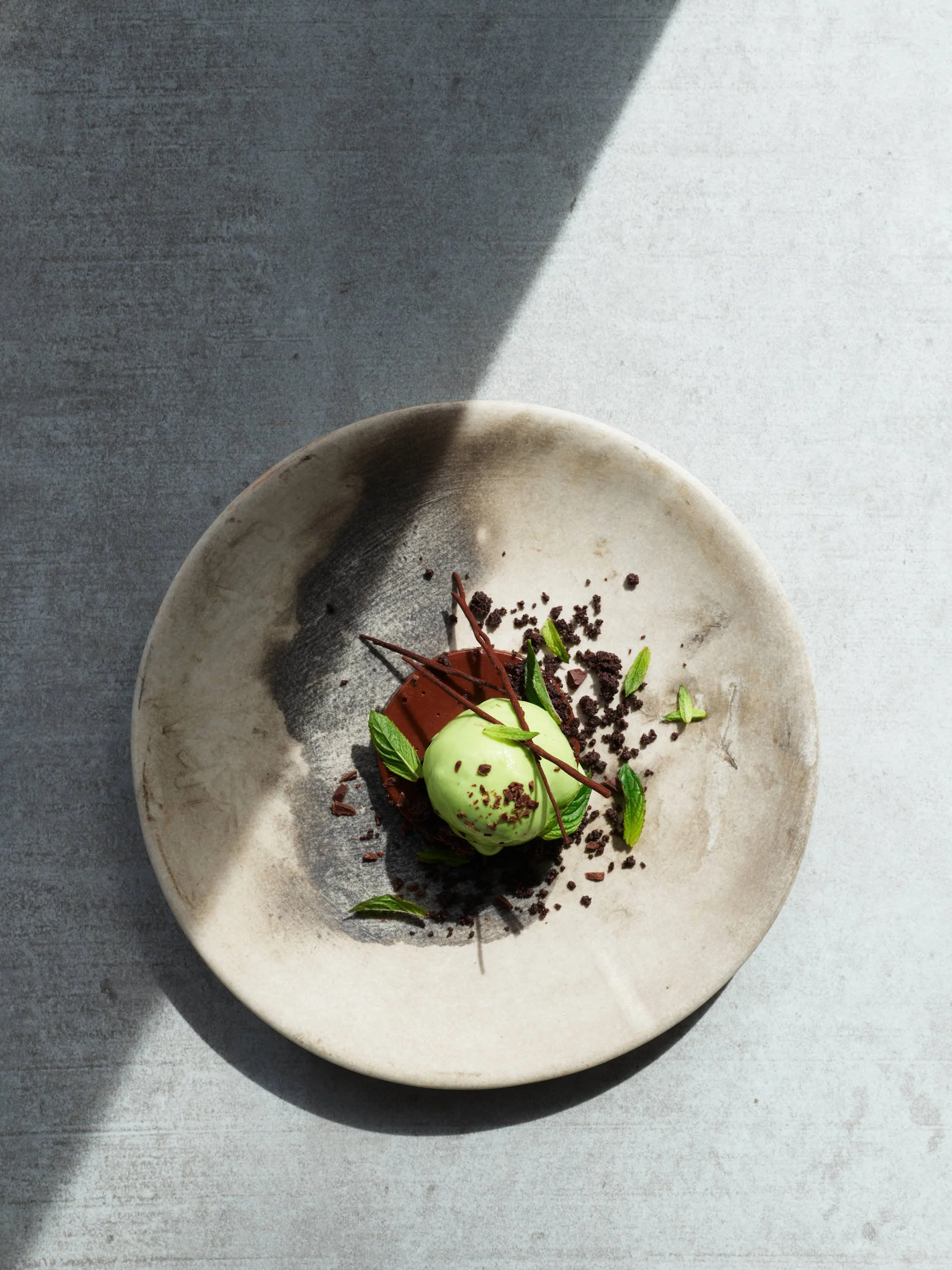 A dessert with a green scoop of ice cream, chocolate garnish, chocolate crumbs, and mint leaves on a rustic ceramic plate with shadow and light.