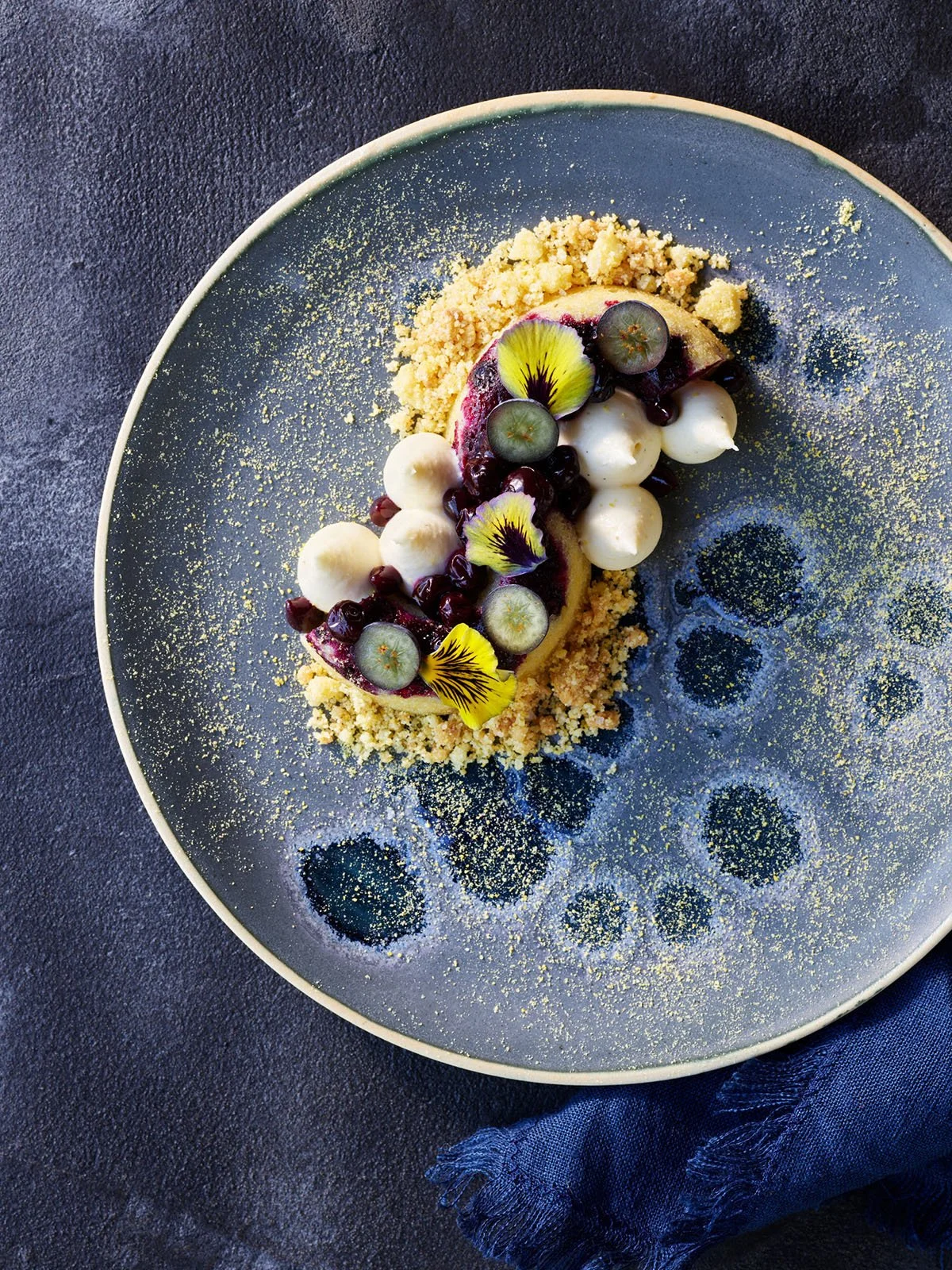 A gourmet dessert with blueberries, edible flowers, white chocolate balls, blueberry sauce, and crumbled crust on a dark plate with a decorative pattern.
