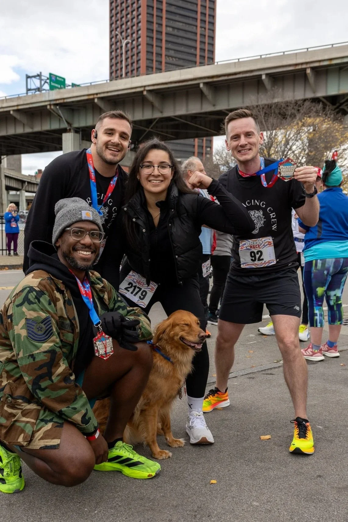 Group of four runners celebrating after finishing a race, with medals around their necks, one holding a medal in hand, and a golden retriever dog with them. They are outdoors under a bridge, with other runners and spectators in the background.