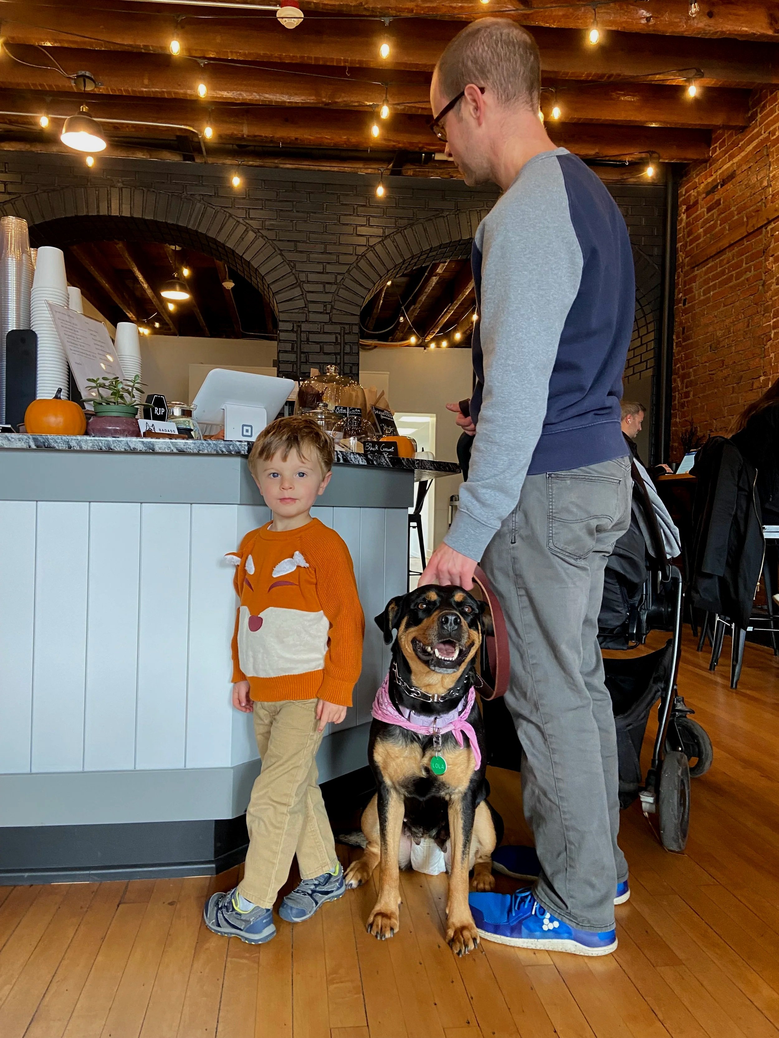 A young boy with a fox-themed sweater standing next to a man with a black and tan dog wearing a pink bandana inside a cafe. The boy has short, light brown hair, beige pants, and sneakers. The man is wearing glasses, a navy and gray sweatshirt, gray pants, and blue sneakers. They are near a counter with a pumpkin, cups, and a cash register, with warm lighting and brick walls.