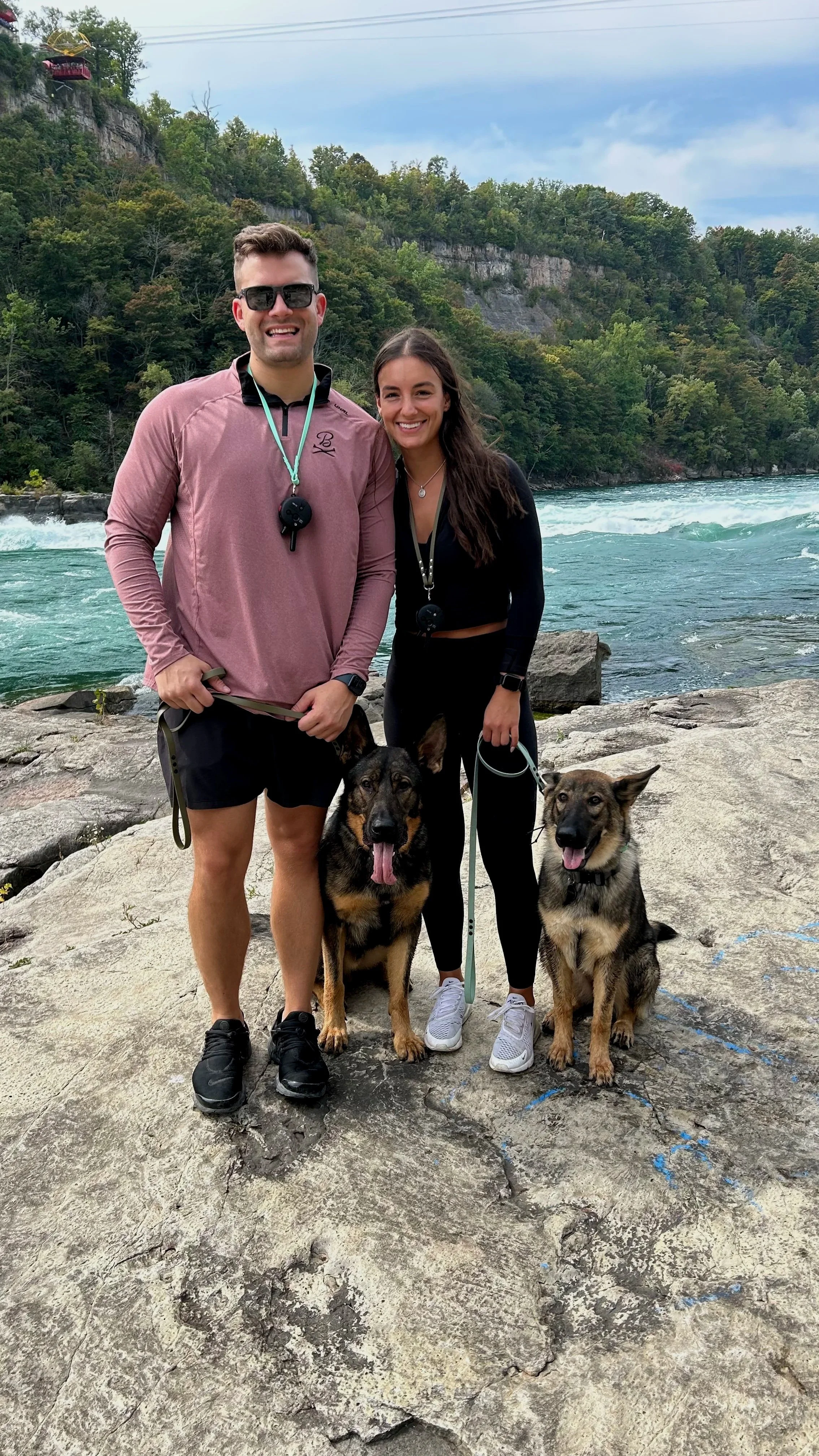 A smiling man and woman standing on rocks by a river with two dogs, surrounded by trees and cliffs.