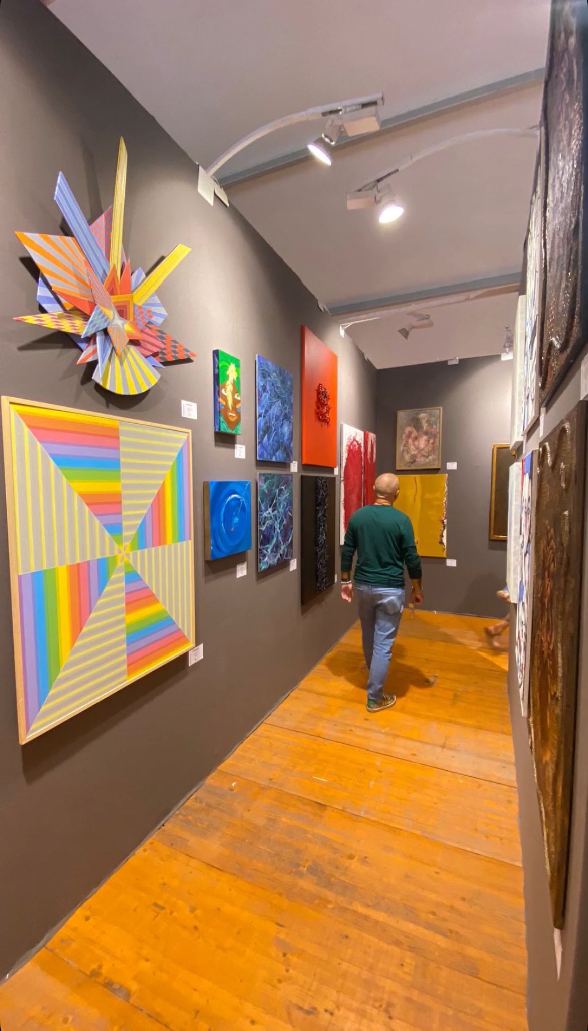 Exposition d'art avec diverses œuvres modernes et abstraites accrochées à un mur gris d'une galerie d'art, un homme marche dans la galerie.